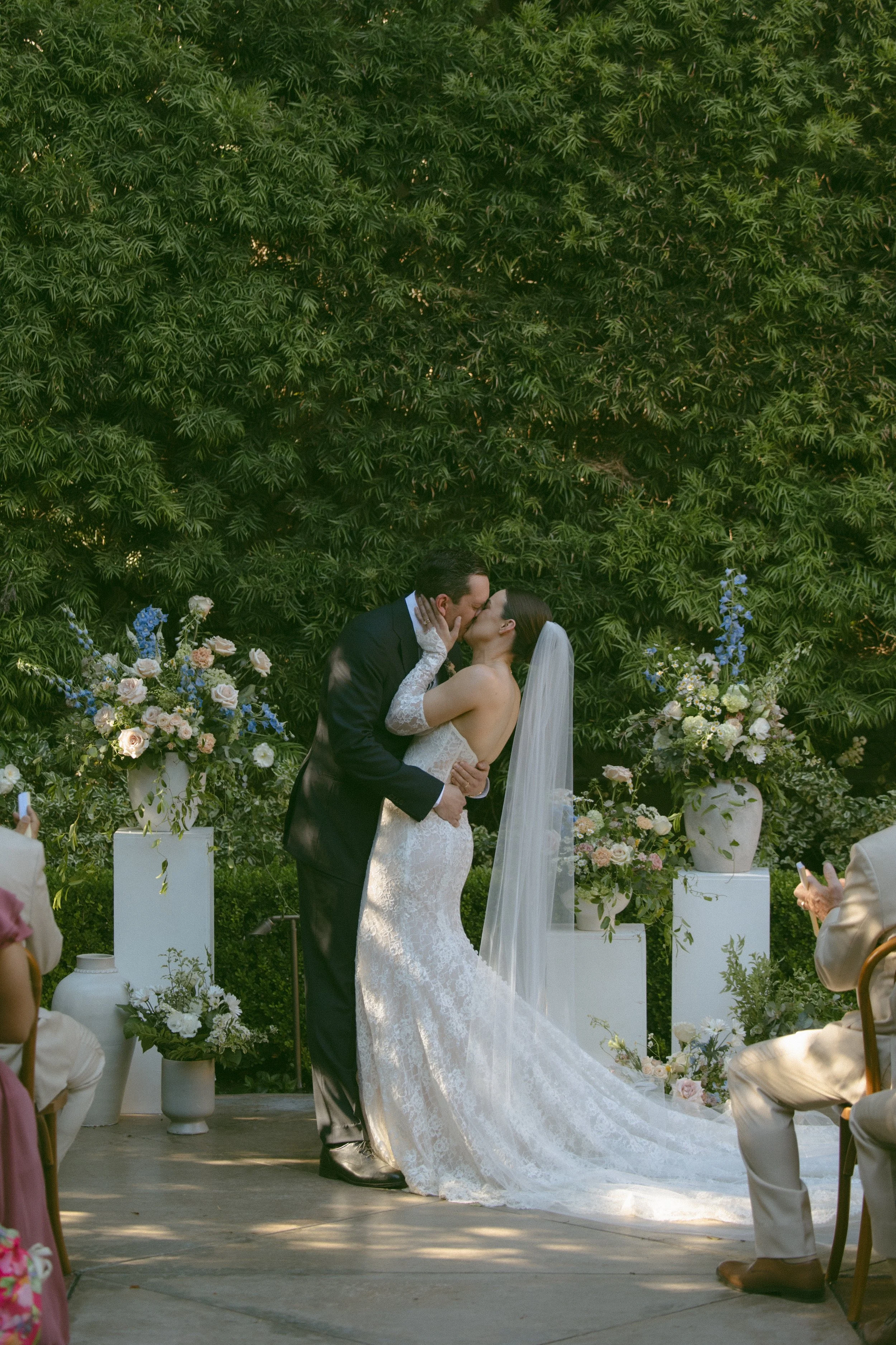 A bride and groom share a kiss at their outdoor wedding ceremony surrounded by floral arrangements and greenery.