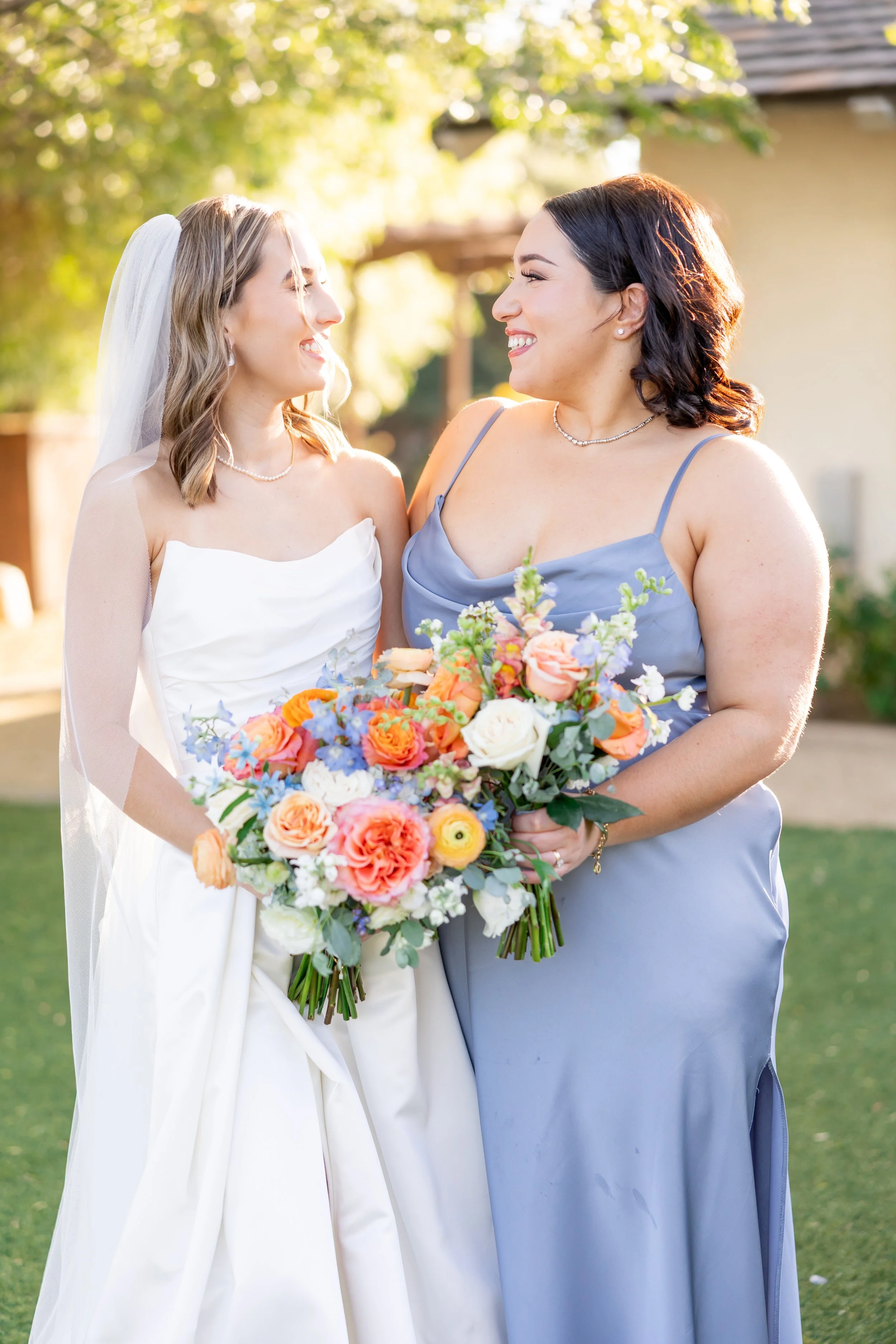 Two women at a wedding, one in a white wedding dress with a veil and the other in a light blue bridesmaid dress, smiling and holding colorful bouquets of flowers outdoors.