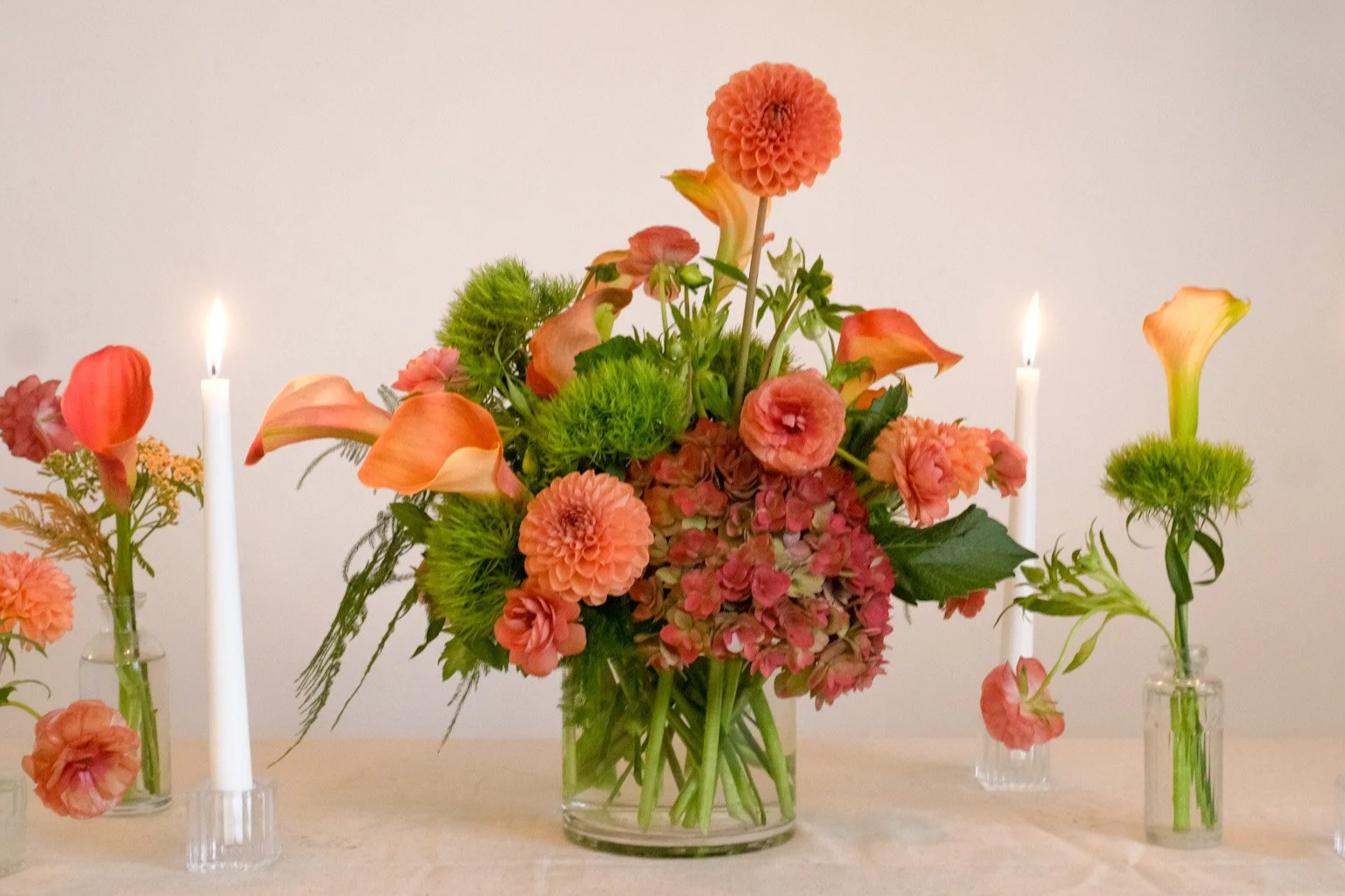 Arrangement of pink and green flowers in a central glass vase, flanked by two tall white candles in small vases on a light-colored table.