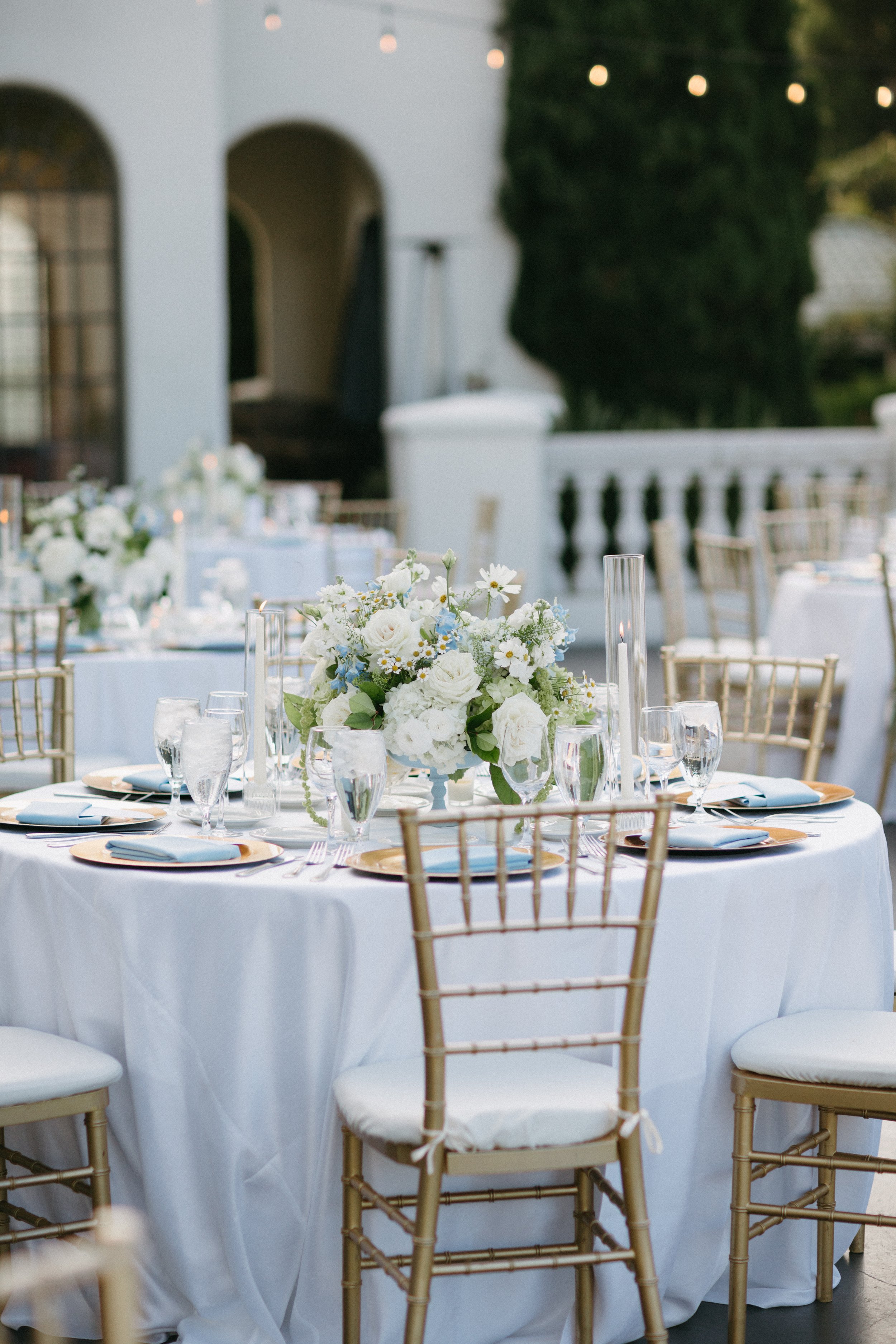 Elegant outdoor wedding reception table with white tablecloth, gold chairs, floral centerpiece with white and blue flowers, and tall glass candle holders, set against a background of a white building and green tree.