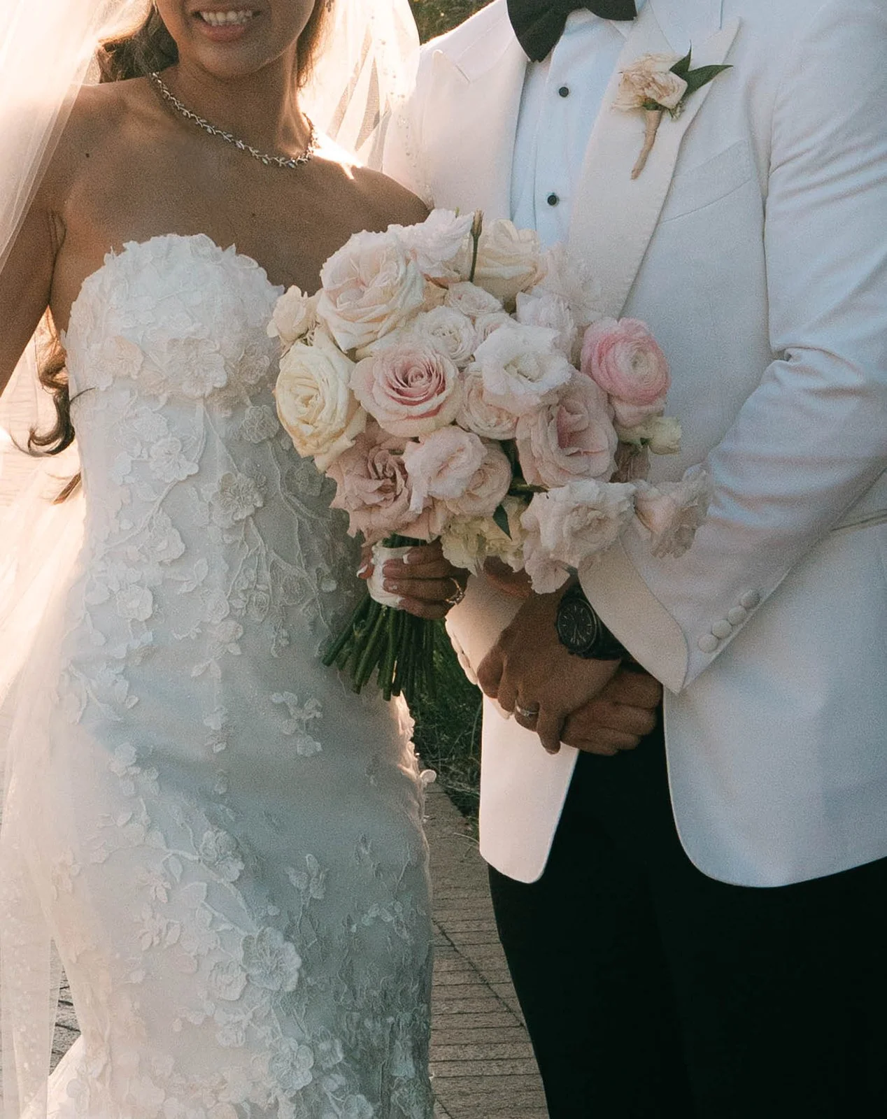 A bride and groom on their wedding day holding a bouquet of light pink and white roses. The bride is wearing a white lace wedding dress and a necklace, and the groom is dressed in a white tuxedo jacket with black trousers and a black bow tie.