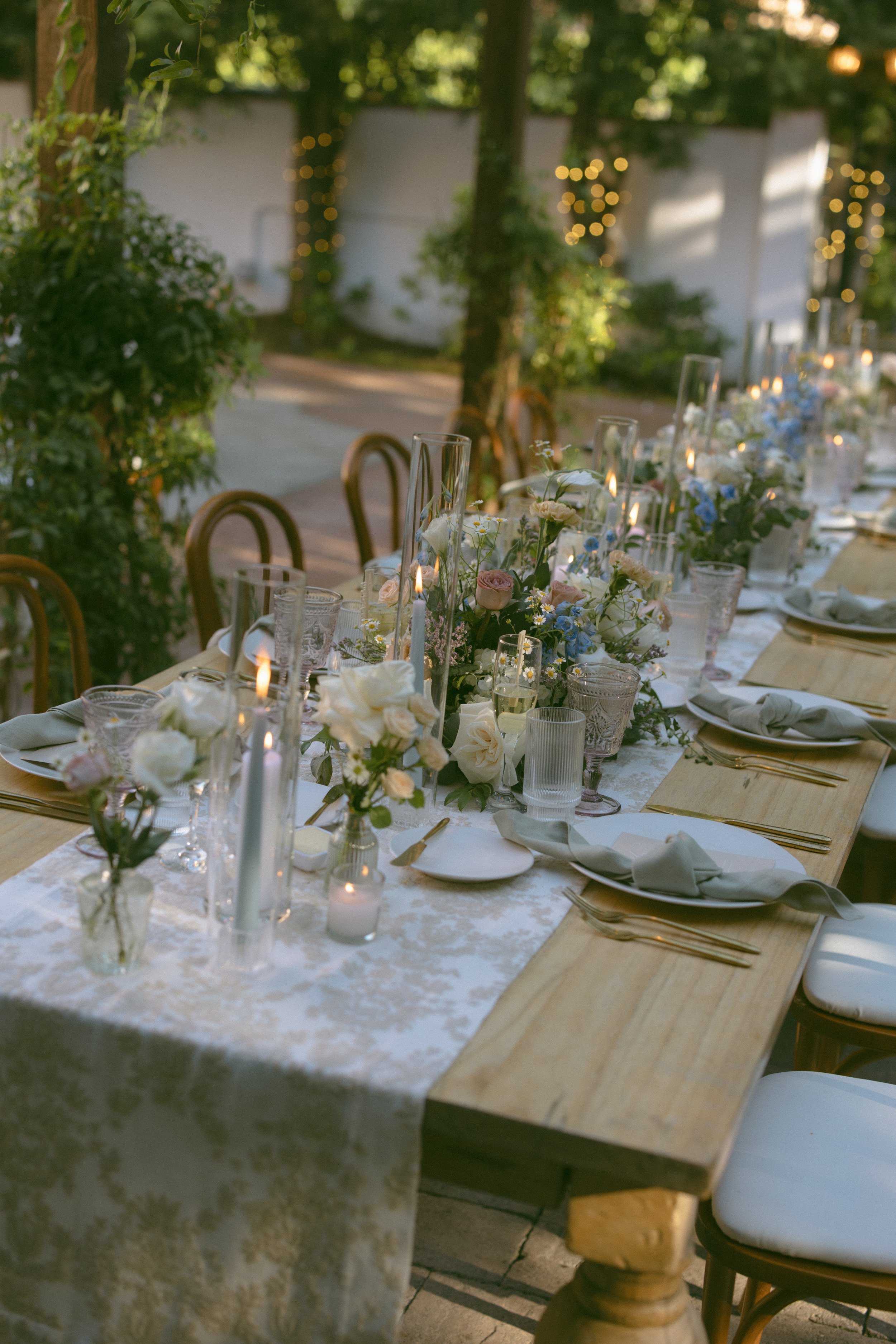 A long outdoor table set for a celebration, decorated with white and pastel flowers, candles, and elegant tableware, with chairs around it and string lights in the background.