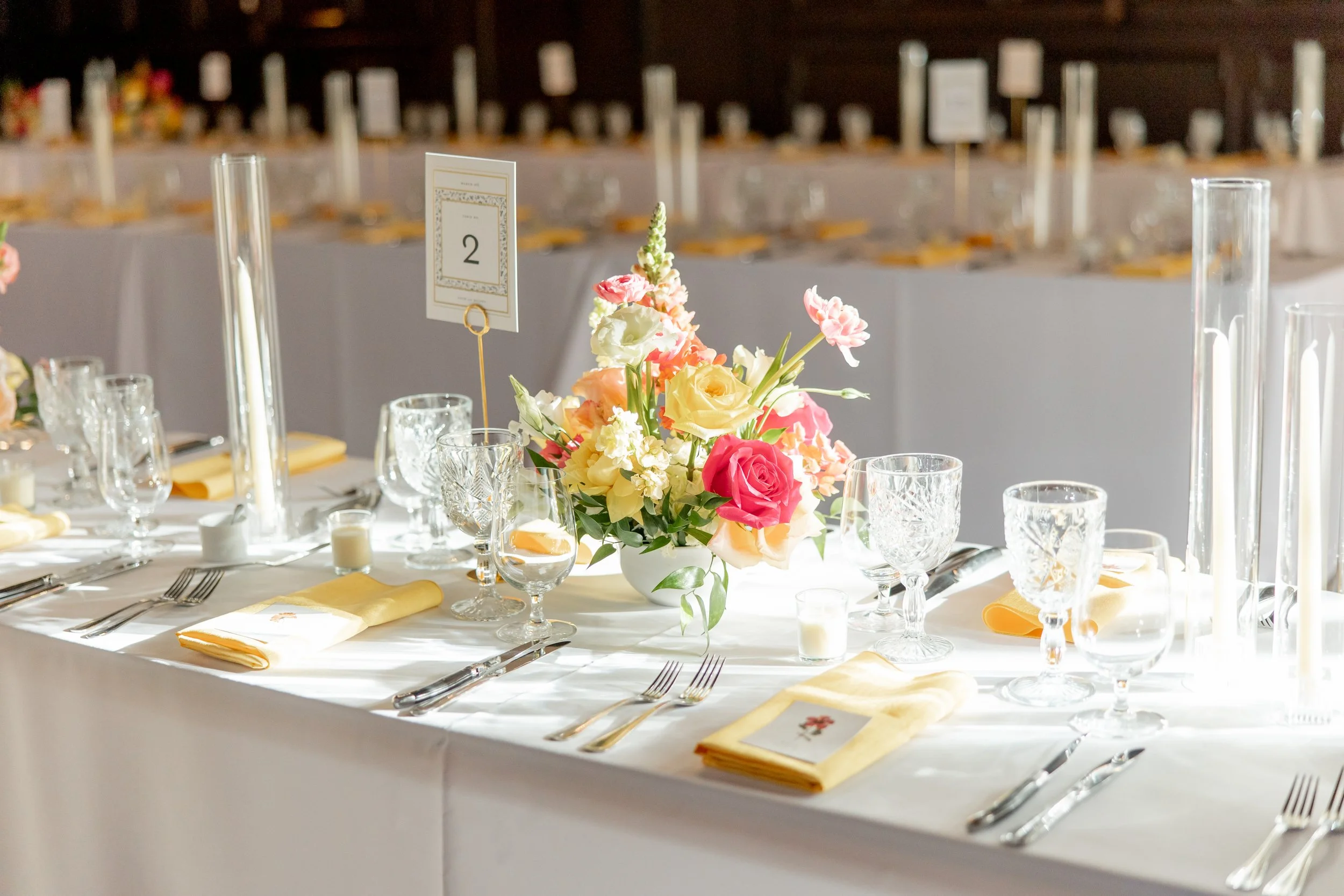 A banquet table decorated with a pink and yellow flower centerpiece, yellow napkins, wine glasses, and candles, set for a formal event.