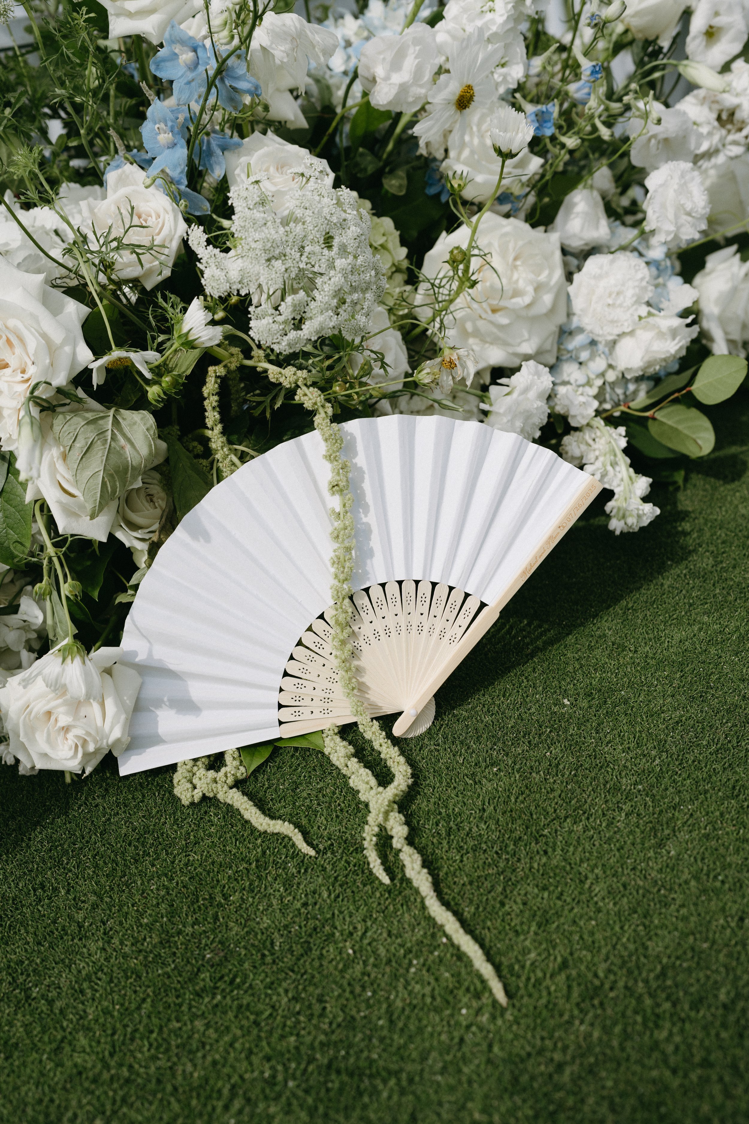 White decorative folding fan resting on green grass with a large arrangement of white flowers and greenery in the background.