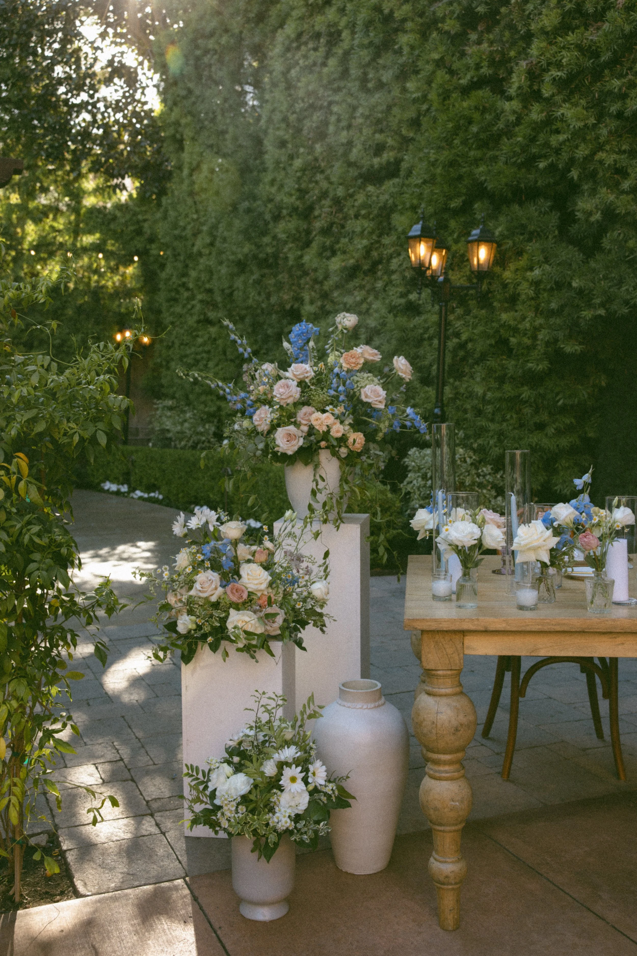 Outdoor event setup with floral arrangements in vases, a wooden table with flower decorations, and a lit street lamp surrounded by lush green trees.