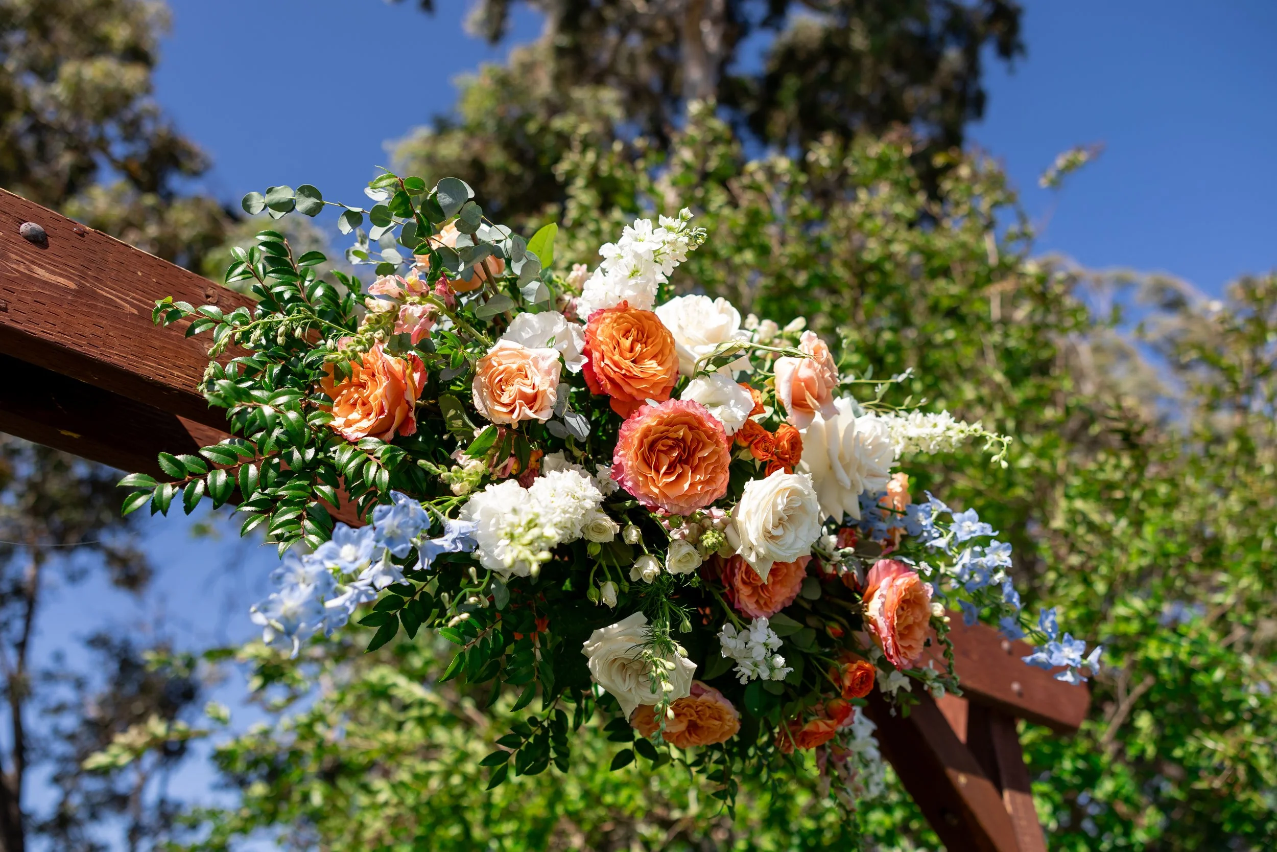 Colorful floral arrangement with roses and greenery attached to a wooden structure, outdoors on a sunny day with trees and a blue sky background.