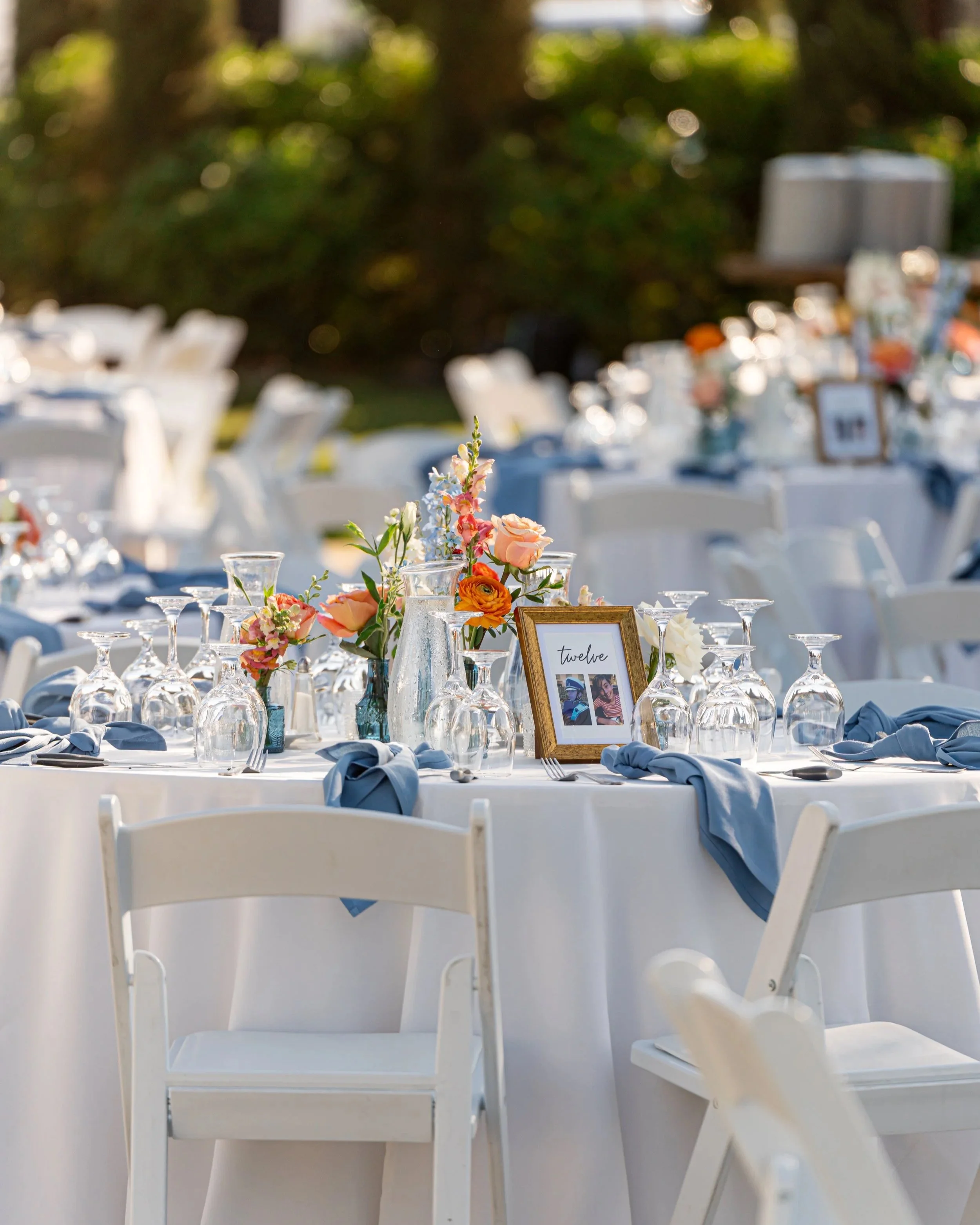 An outdoor event setup with tables decorated with floral centerpieces, glassware, and blue napkins; a framed table number with photos; white chairs; sunlight and greenery in the background.