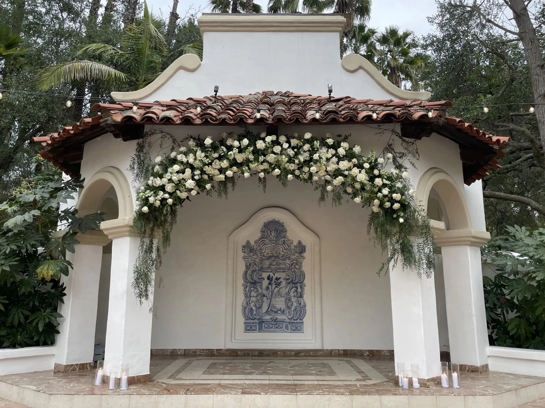 Small, white chapel with a red-tiled roof decorated with white flowers and greenery, with candles at the base of the pillars and a mural of a religious figure on the back wall.