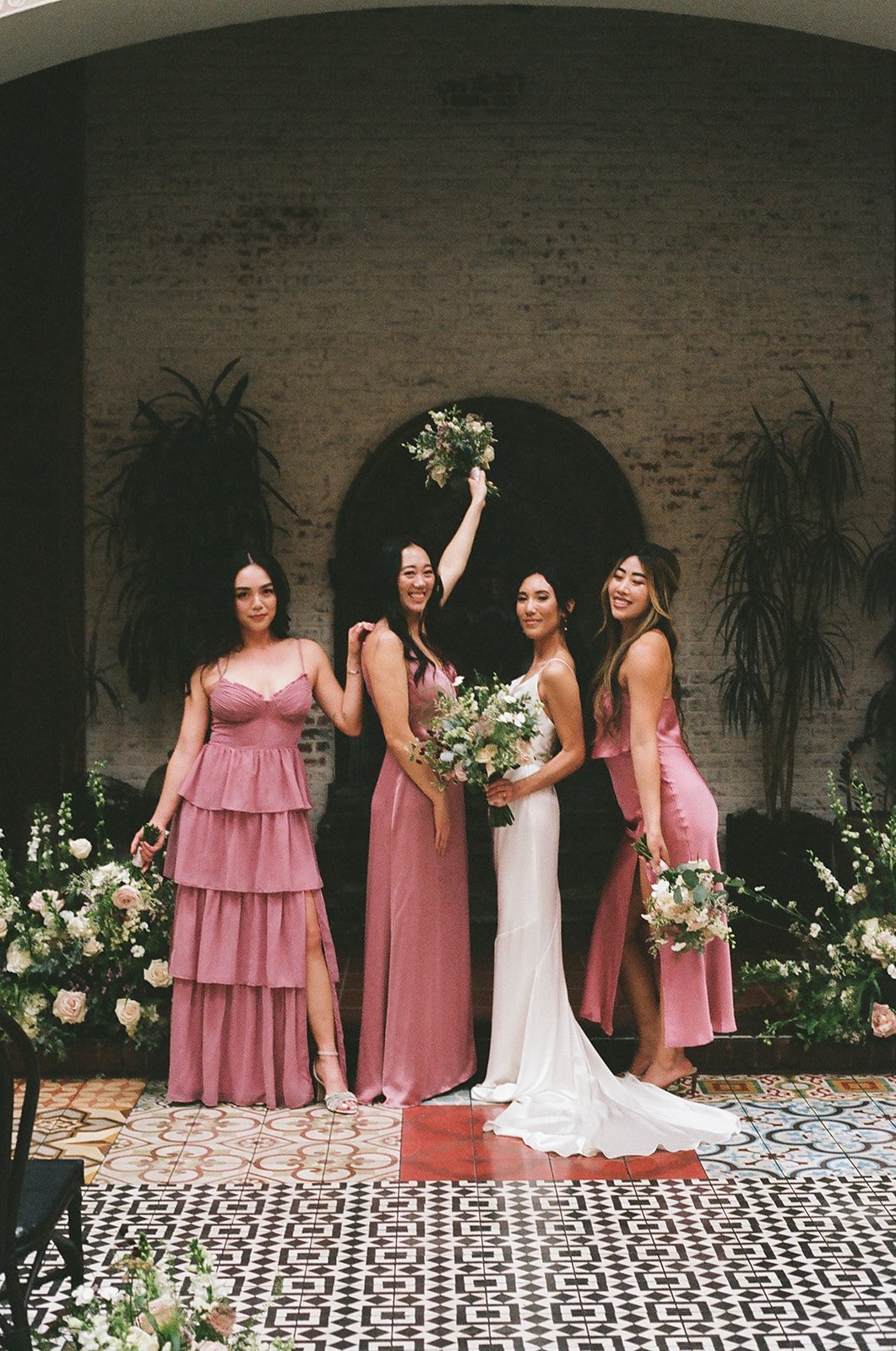 Four women in pink dresses, with the bride in white, in a wedding setting with flowers and plants, posing for a photo.