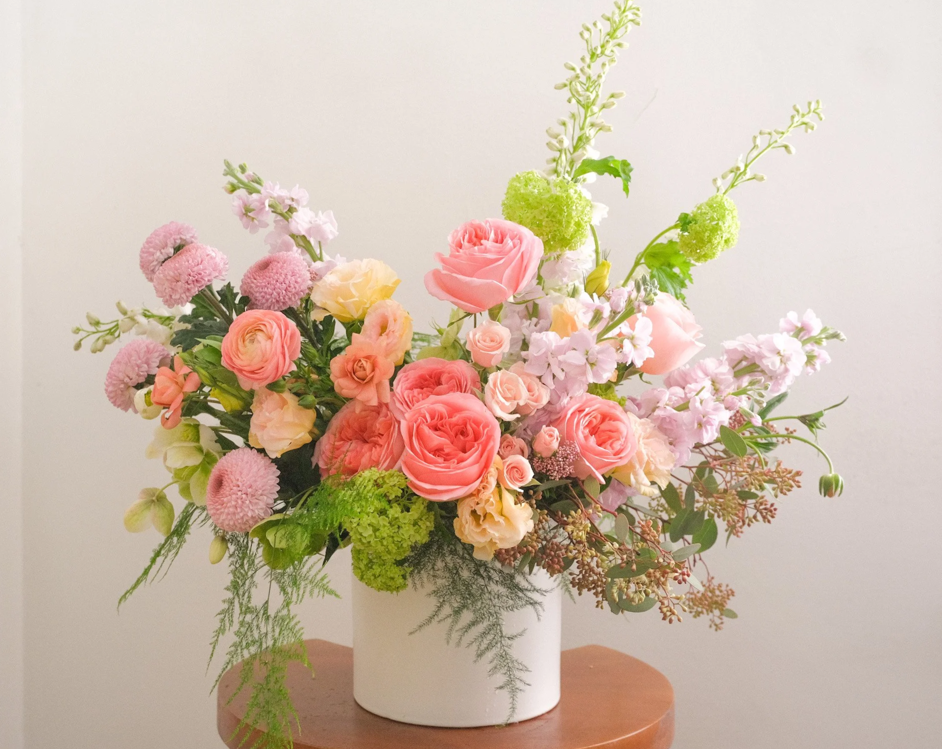 A colorful bouquet of pink, peach, and white roses, along with other pastel-colored flowers and greenery in a white vase on a wooden surface.