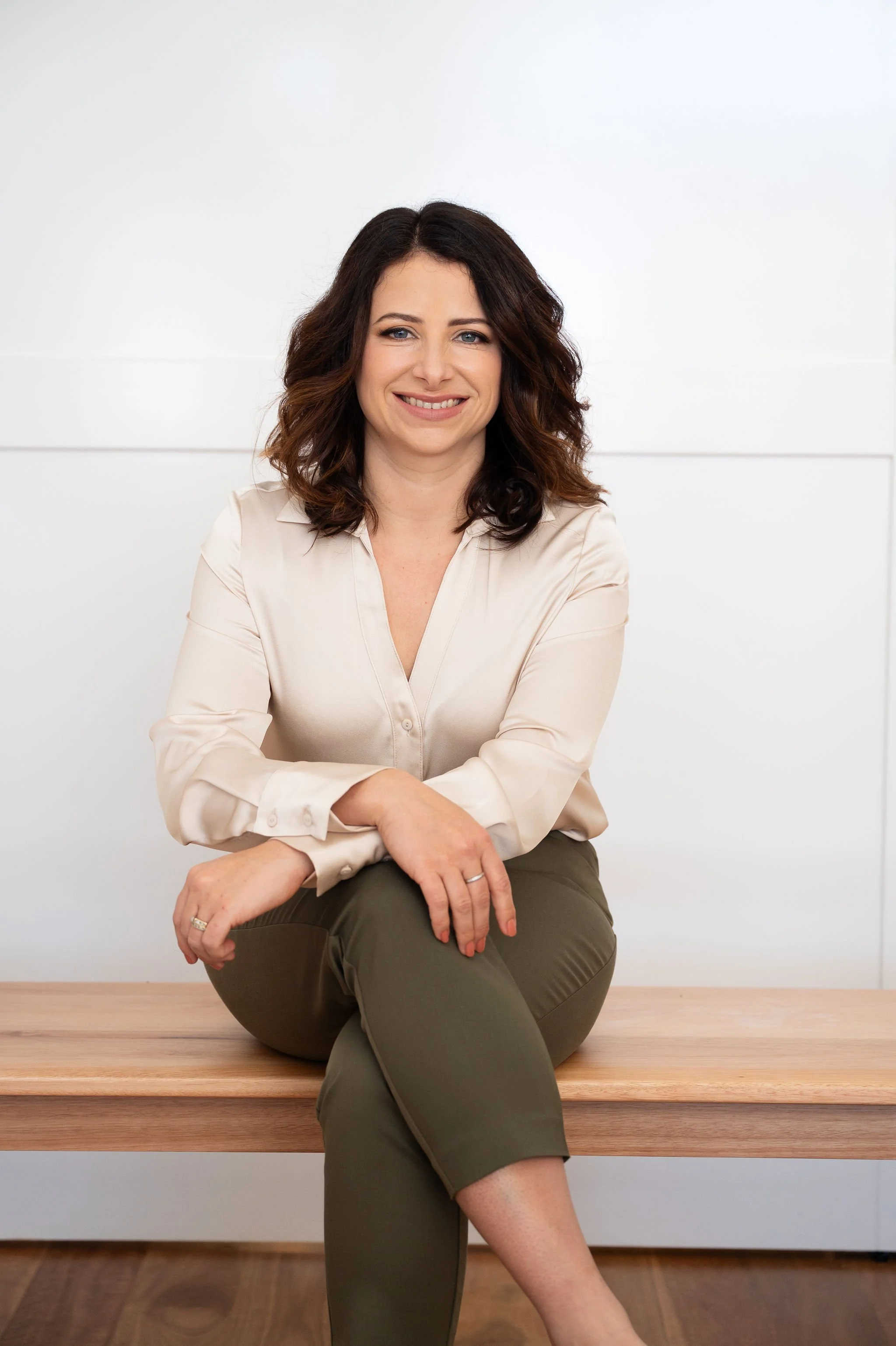 A woman with dark brown, curly hair and light skin sits on a wooden bench against a plain white background, smiling at the camera. She is wearing a cream-colored silk blouse and olive green pants.