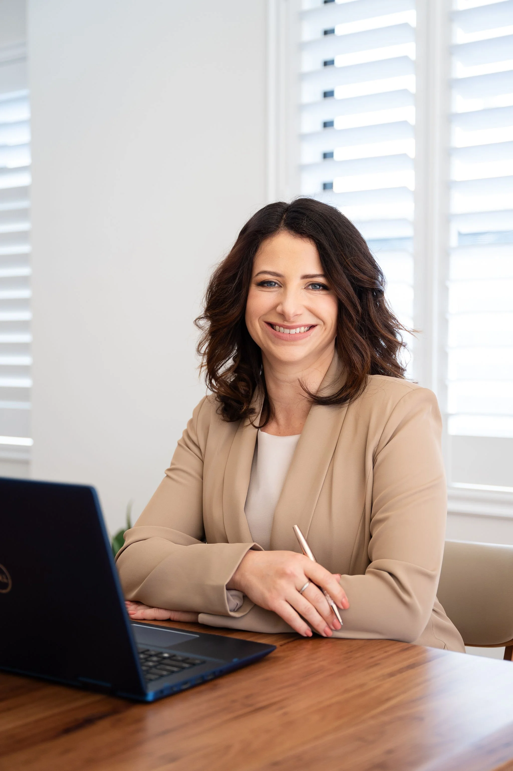 A smiling woman in a beige blazer sitting at a desk with a laptop.