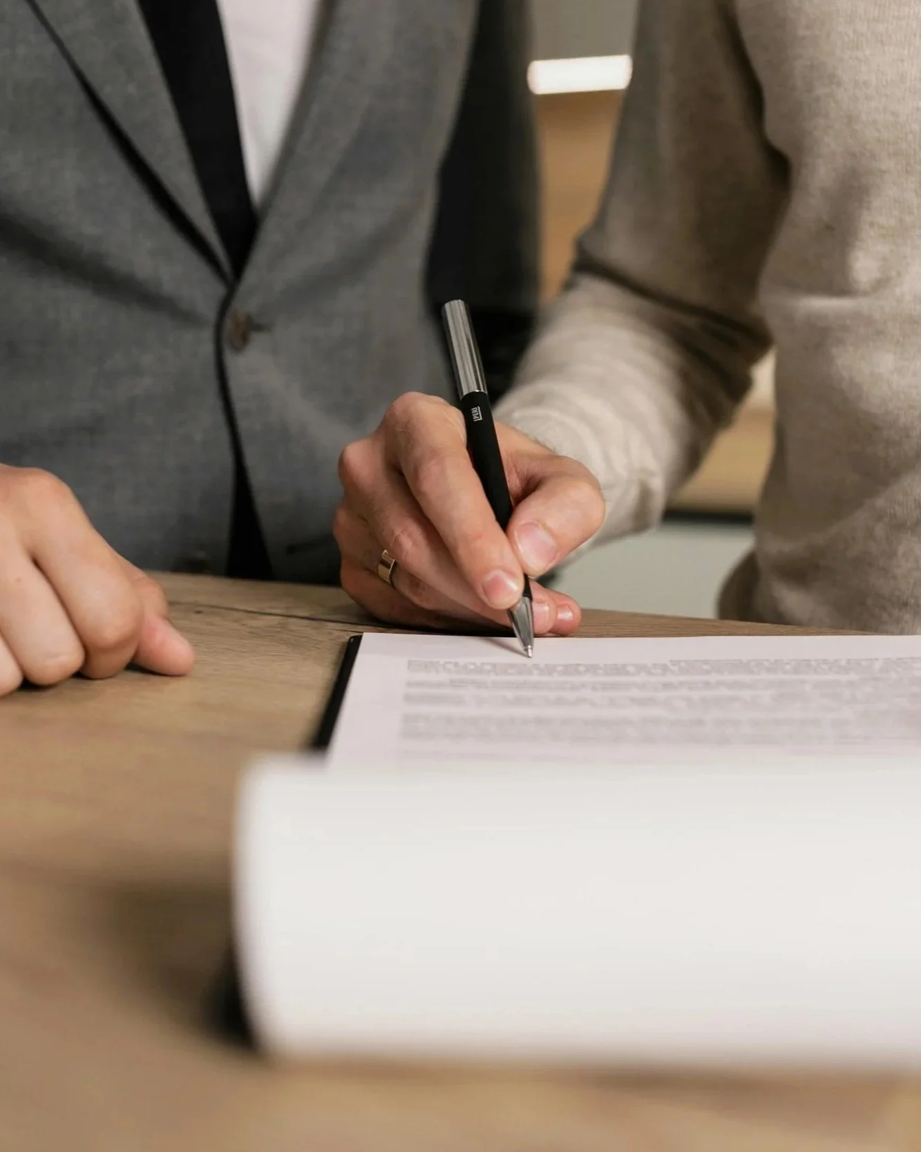 Two people signing a document on a wooden table, one person wearing a gray blazer and the other in a light gray sweater.