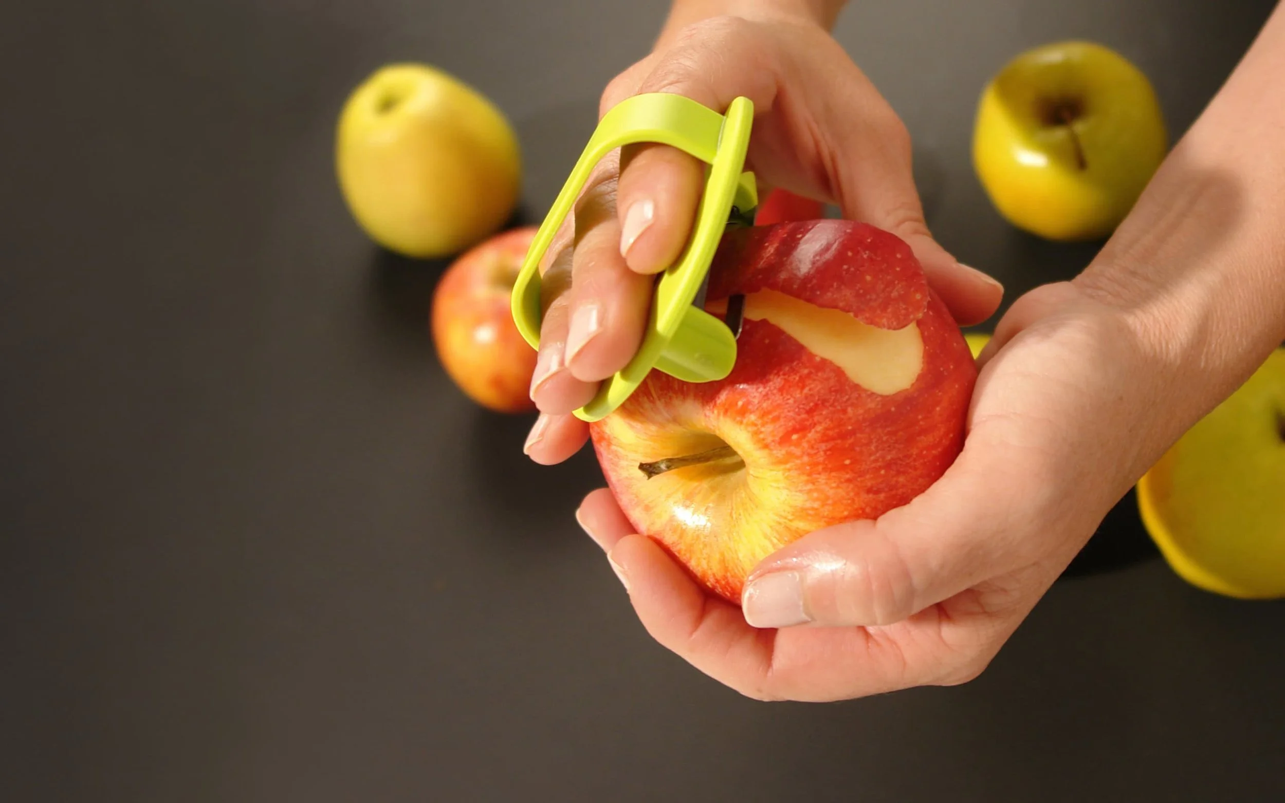 Person schält einen roten Apfel mit einem grünen Schäler, um das Fruchtfleisch sichtbar zu machen, während andere grüne und rote Äpfel auf einem schwarzen Hintergrund liegen.