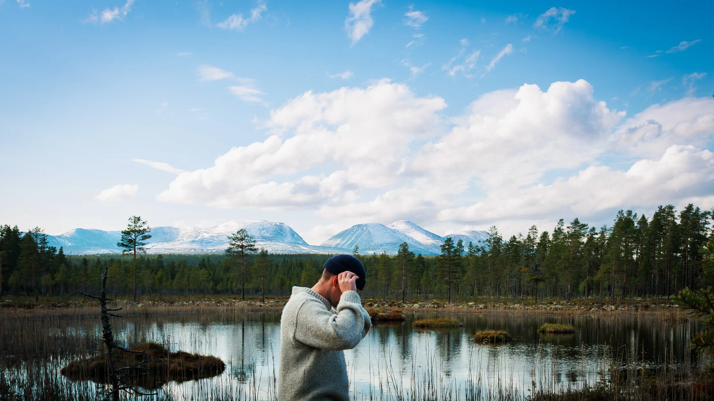 A man wearing a gray sweater and a dark beanie hat standing near a wetland with tall grasses, trees, and snow-capped mountains in the background under a partly cloudy sky.