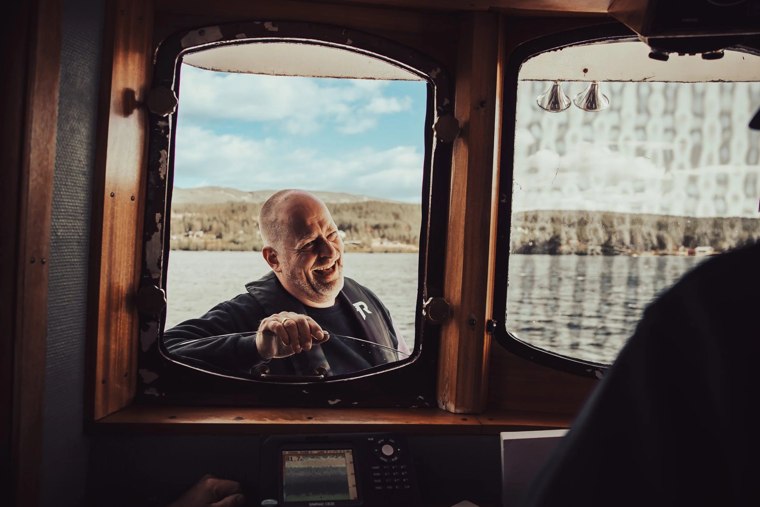 A man smiling and leaning out of a boat's window towards the camera, with a body of water and hills in the background.