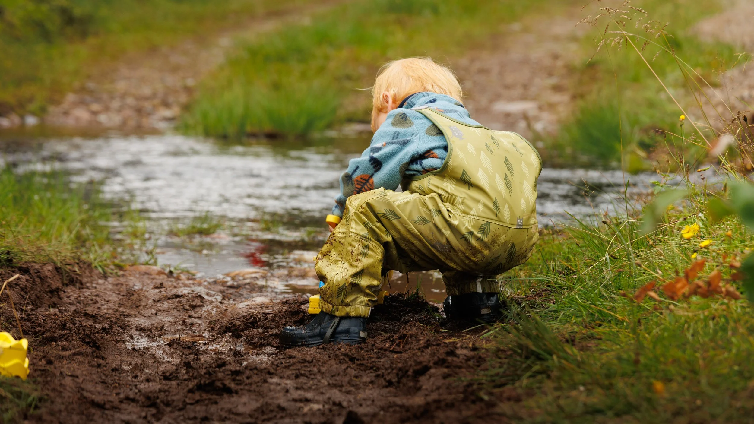 A young child with blond hair, wearing a colorful fleece jacket and waterproof boots, crouches by a small stream in a natural outdoor setting.