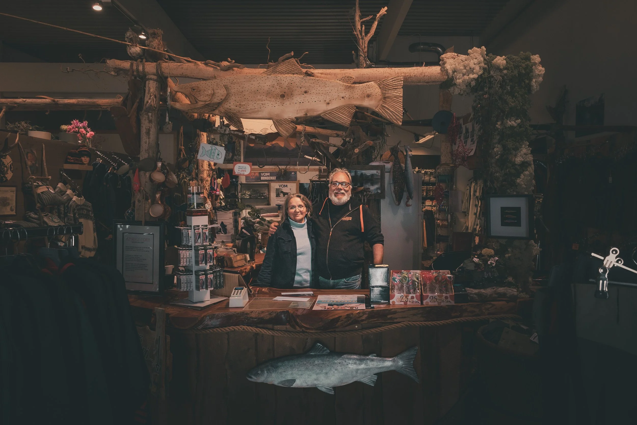 Two people, a woman and a man, standing behind a wooden counter inside a shop with fishing and outdoor gear, with a large mounted fish on the ceiling and a smaller fish on the front of the counter.