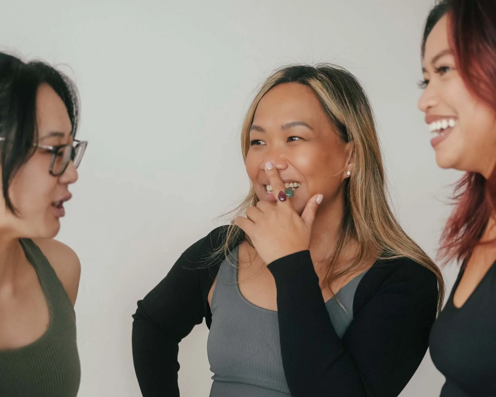 Three women talking and laughing together, one woman in the middle touching her nose with her hand, the woman on the right wearing a dark top, and the woman on the left wearing glasses and a sleeveless top.