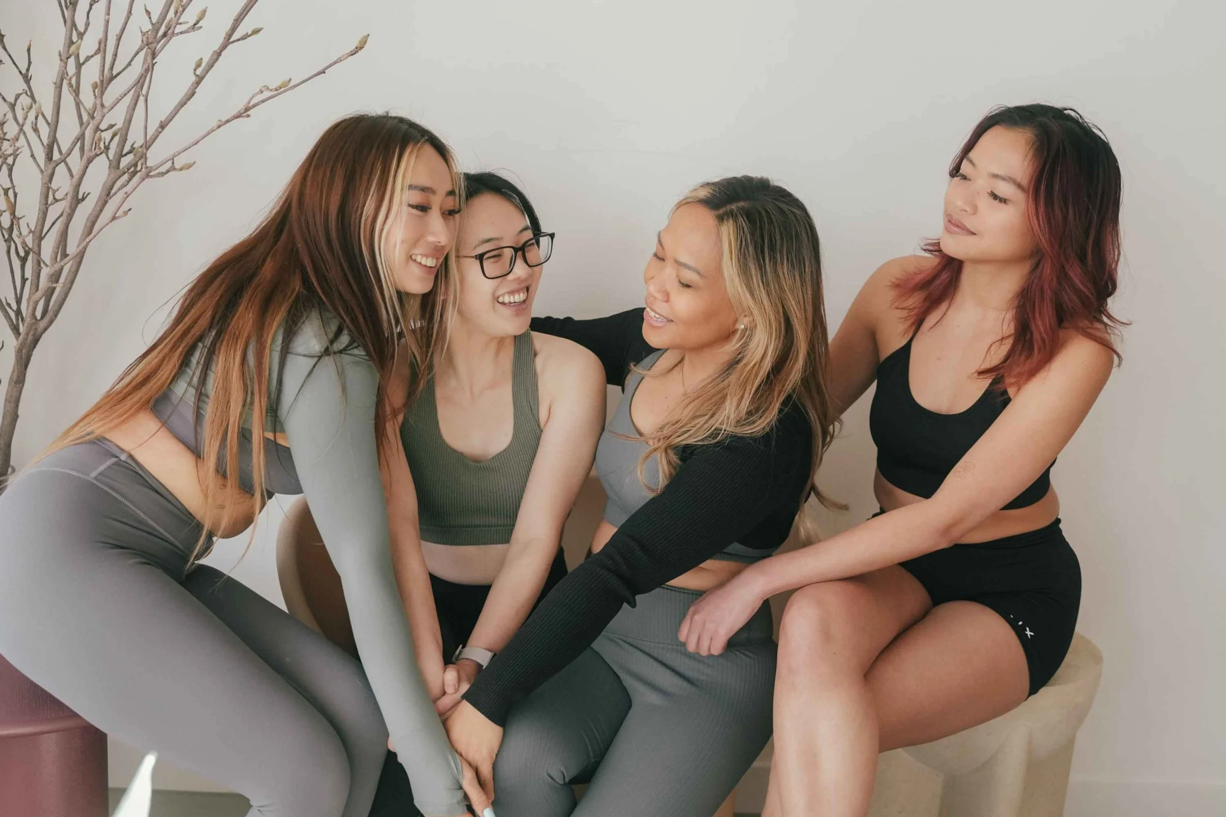 Four women are sitting close together, smiling, and engaging playfully in a group activity indoors.