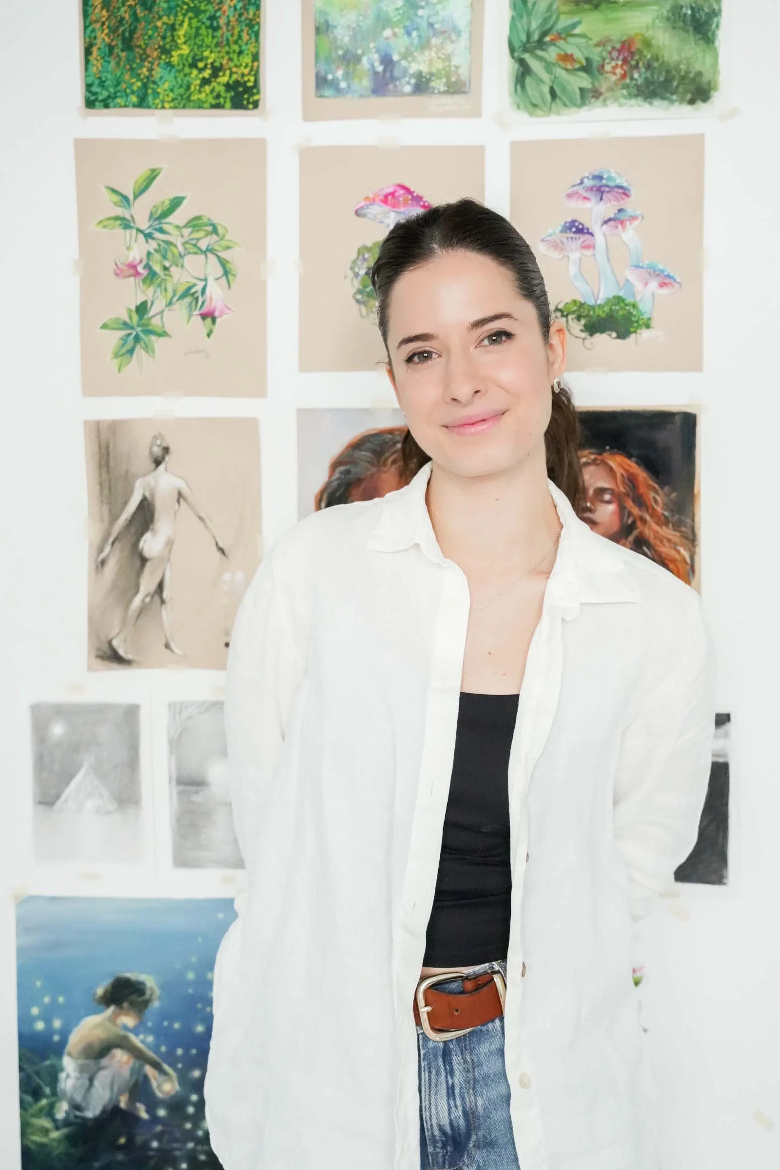 A young woman with dark hair tied back, wearing a white shirt over a black top and blue jeans, standing in front of a wall displaying various colorful artworks.