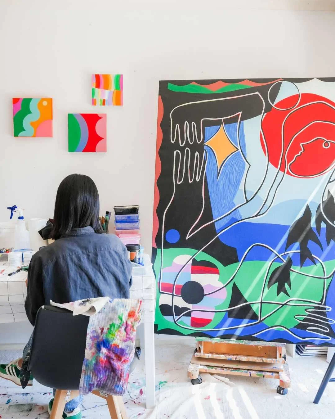 An artist working on a colorful abstract mural in a studio. The mural features bold lines and vibrant colors including red, blue, green, and black. Several smaller abstract paintings hang on the white wall behind the artist.