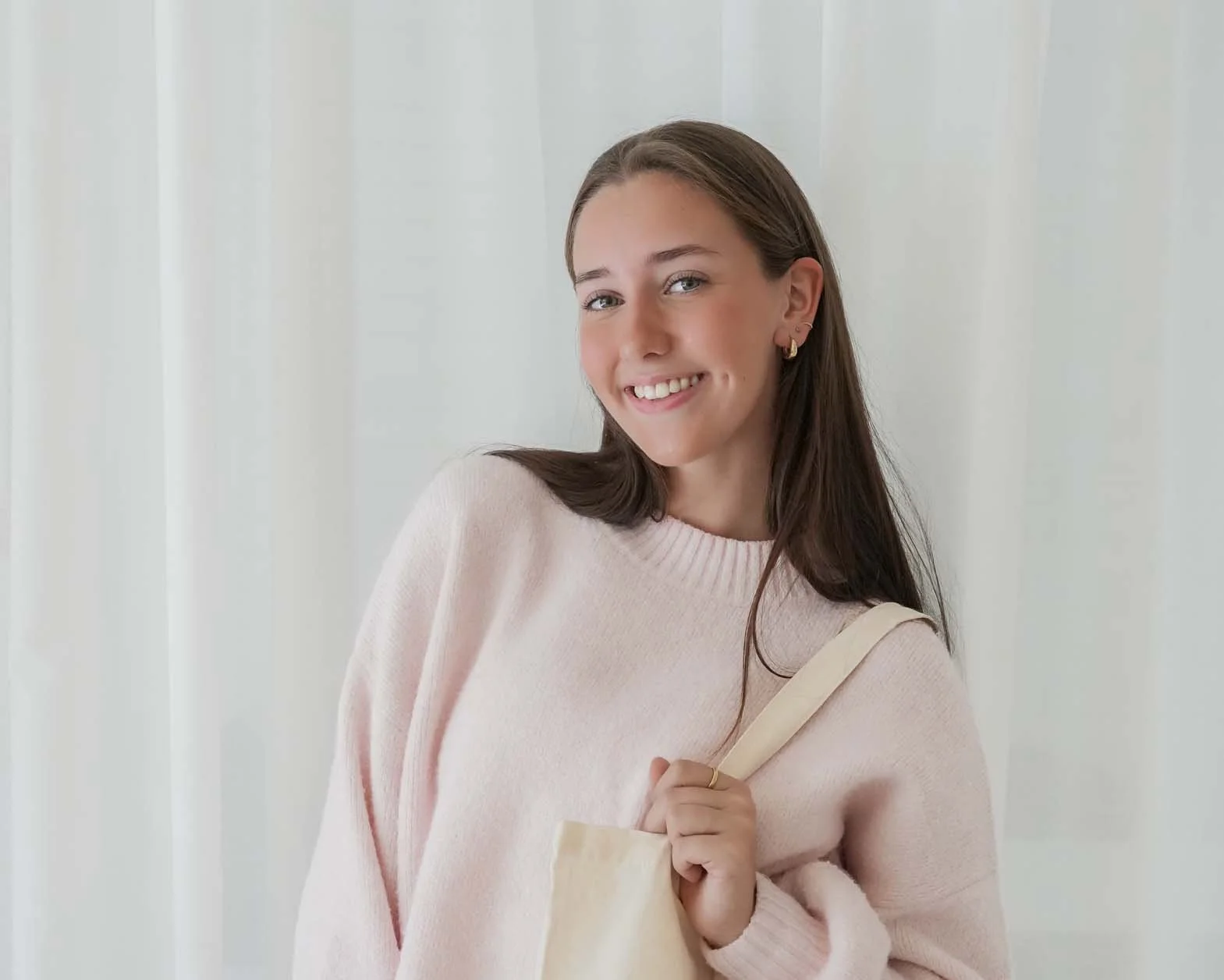 A young woman with long brown hair smiling and holding a beige tote bag, wearing a light pink sweater, standing in front of a white wall.