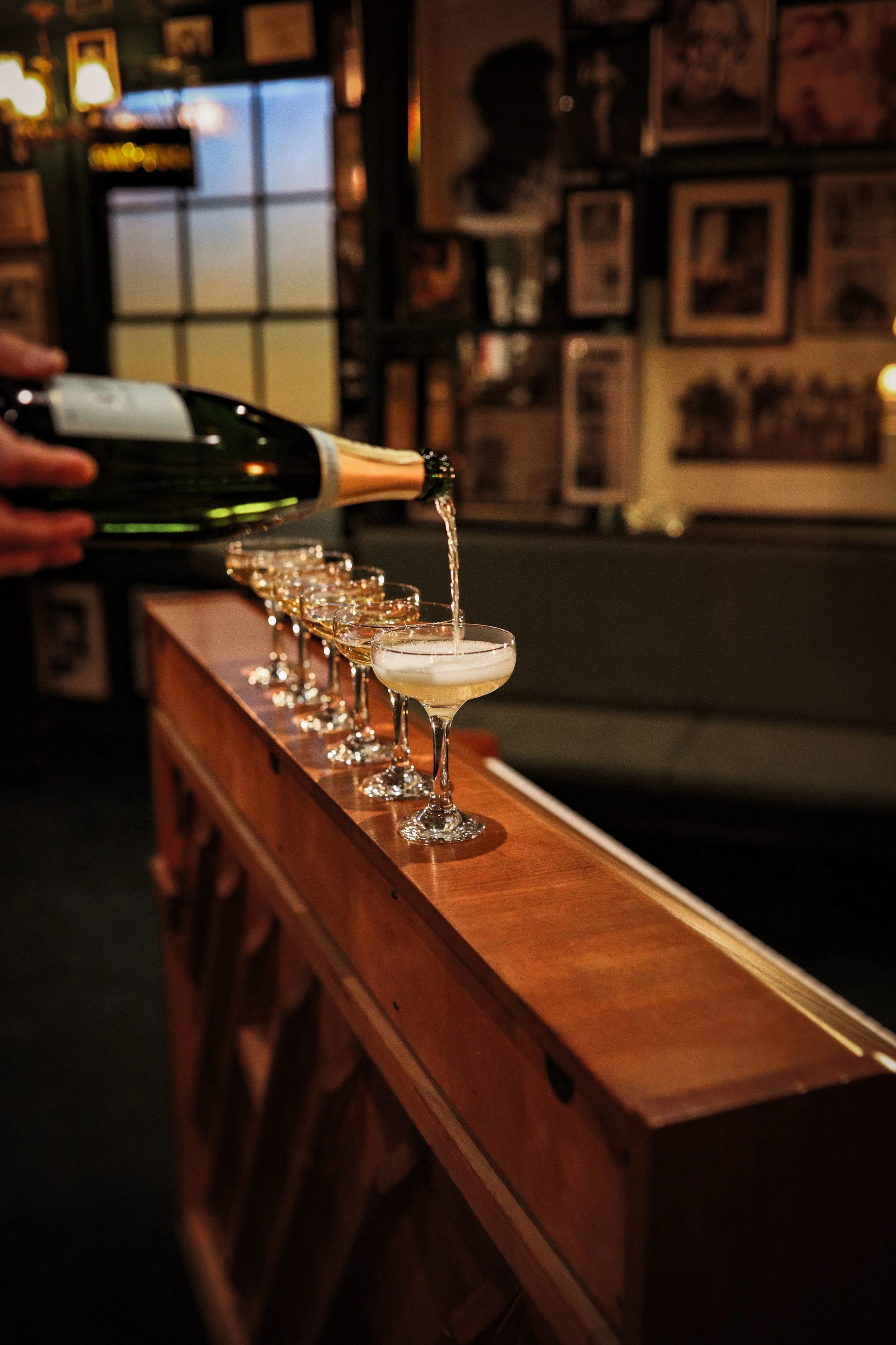 Person pouring champagne into a row of coupe glasses on a wooden bar in a dimly lit bar or restaurant.