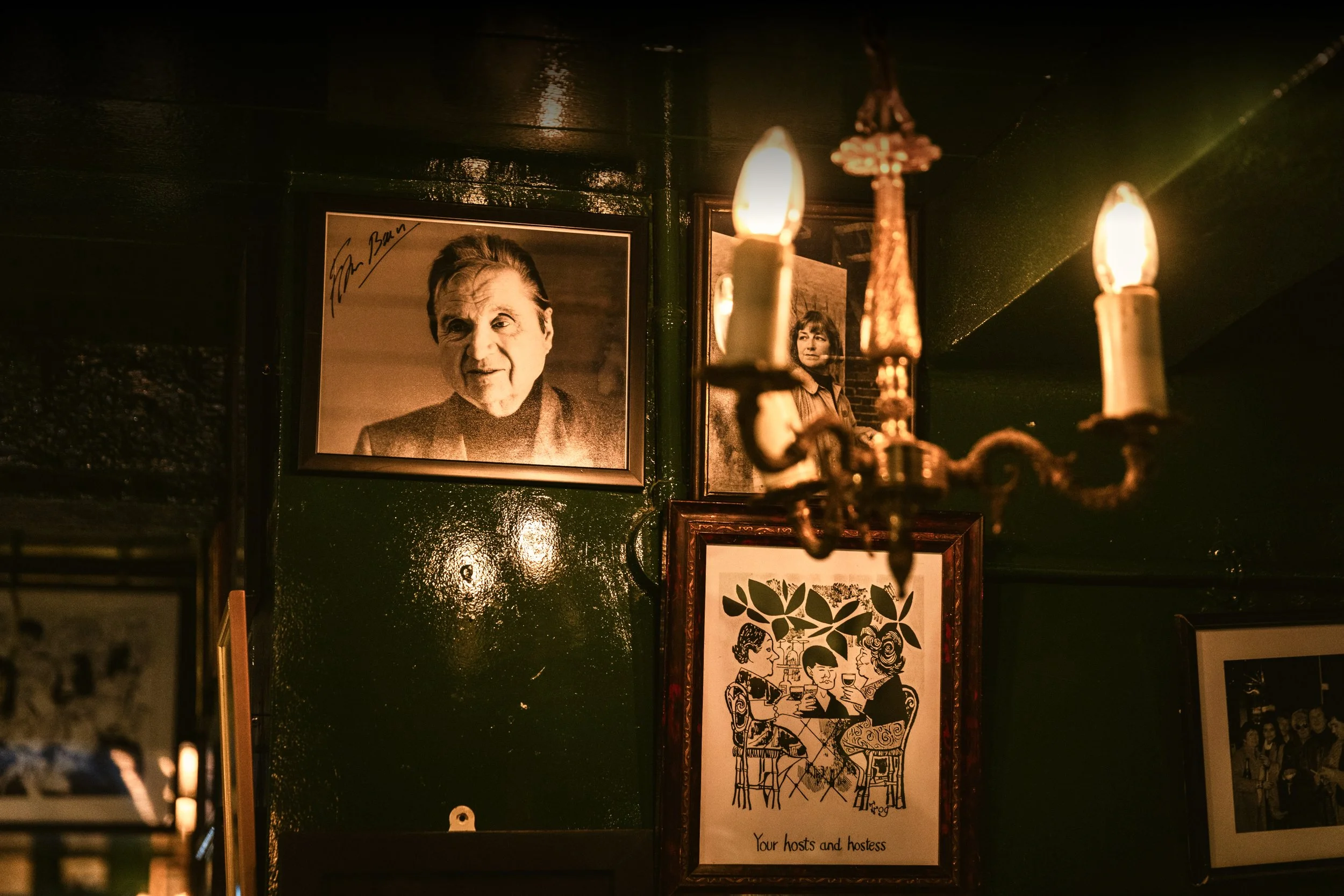 Dimly lit wall with framed photographs, one of a man signing, and a colorful illustration of people dining, under a gold chandelier with lit candles.