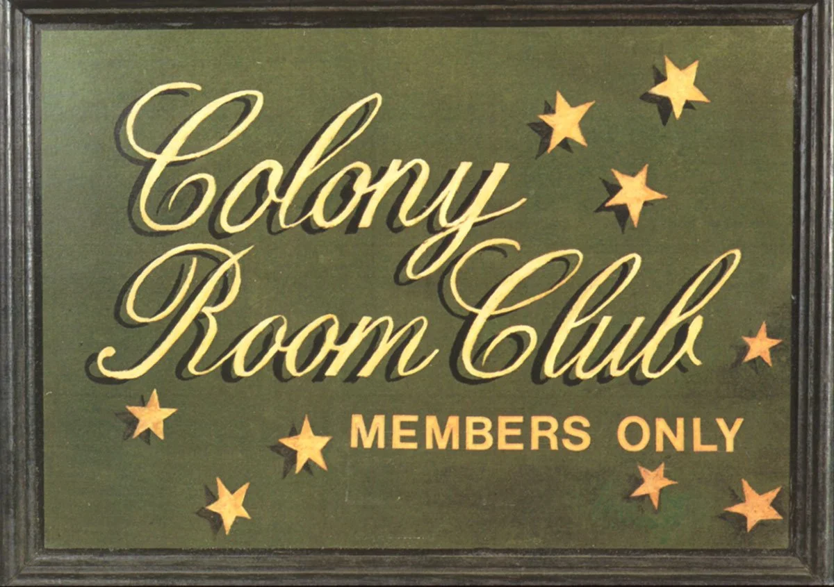 A sign with a dark green background and black frame that reads 'Colony Room Club' in yellow cursive lettering and 'MEMBERS ONLY' in yellow uppercase letters. Decorative yellow stars surround the text.