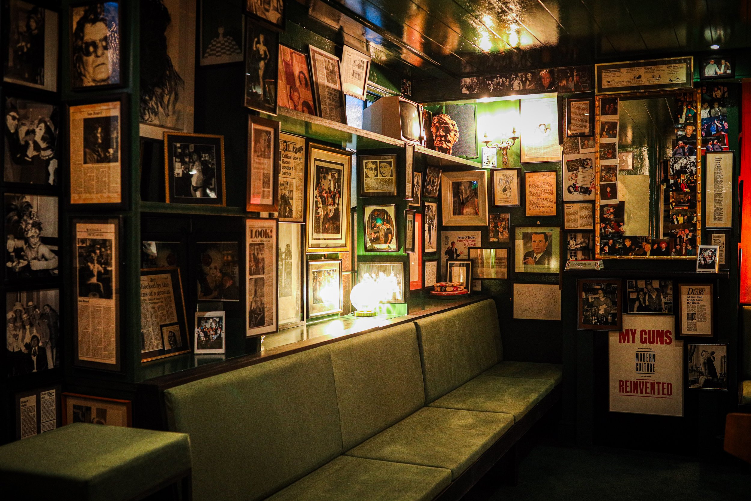 A dimly lit interior of a cafe or bar with dark walls decorated with numerous framed photographs, clippings, and artwork. There is a long green cushioned bench along the wall, and a small table with a lit lamp in the background.