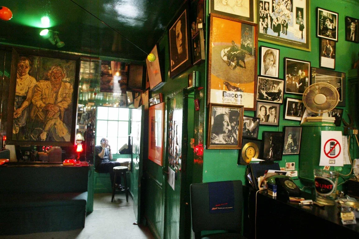 Interior of a bar or cafe with green walls decorated with framed photos and artwork, including black-and-white and colored pictures, a fan, a sign, and a person sitting by a window.