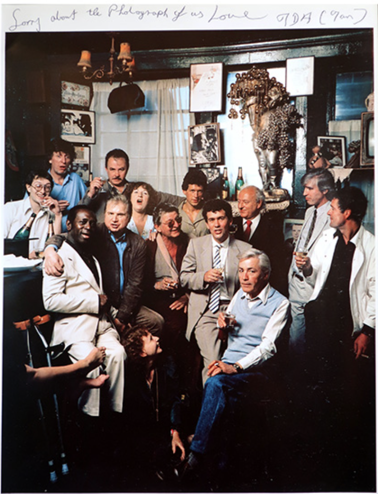 A group of people at a party in a cozy, art-deco style room with dark wooden walls, framed pictures, and decorative objects. Some are sitting, some standing, holding drinks, and smiling for the photo.
