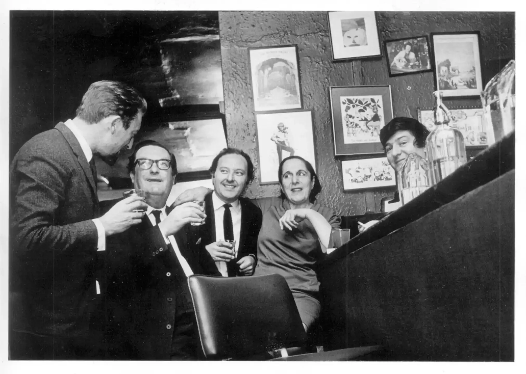 A group of five adults at a bar or pub, smiling and holding drinks, with artwork and photos on the wall behind them, including a window.