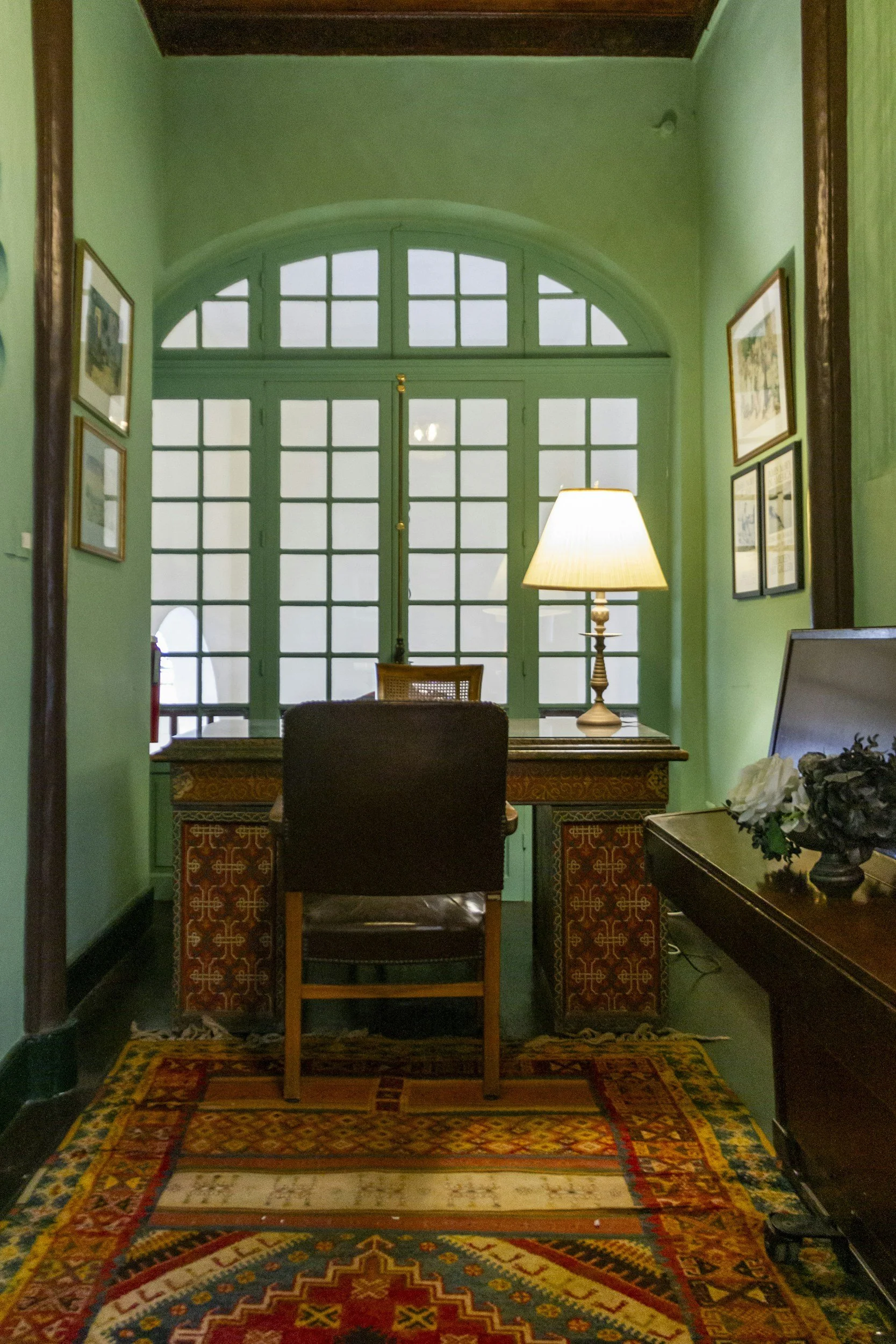 Interior room with green walls, a large arched window with multiple panes, a wooden desk with intricate carvings, a black chair in front, a lit table lamp, framed pictures on the wall, and a colorful patterned rug.
