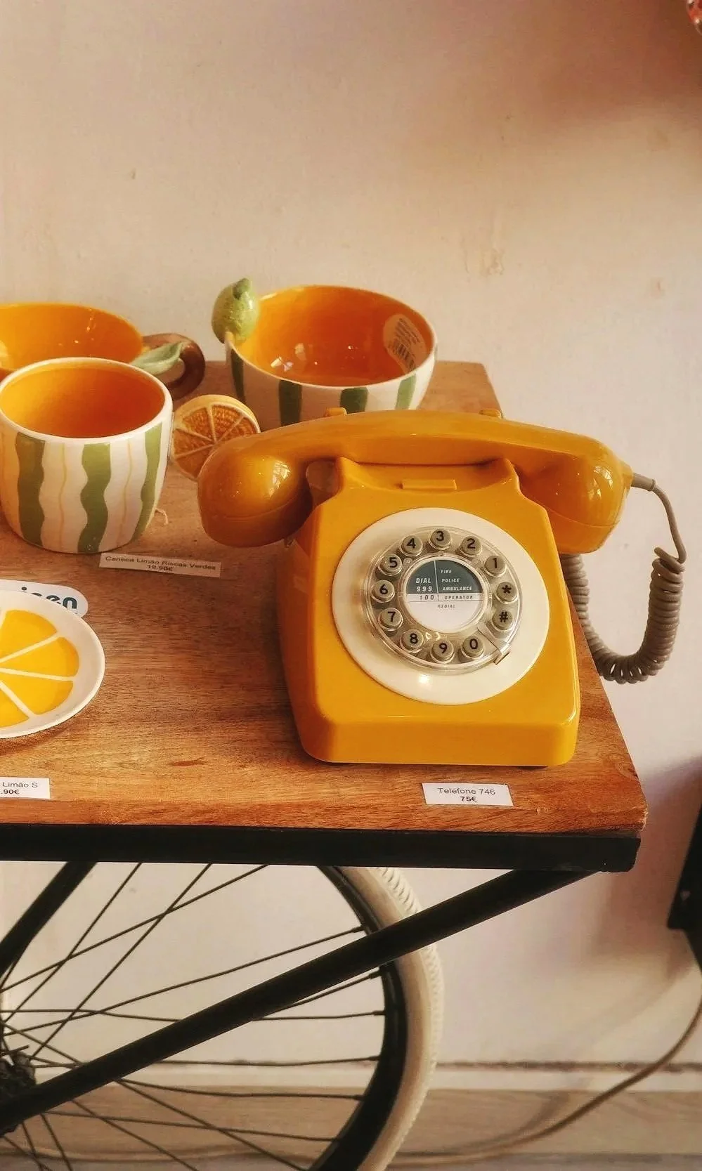 A vintage yellow rotary telephone on a wooden table surrounded by yellow and green cups and a small lemon plate.