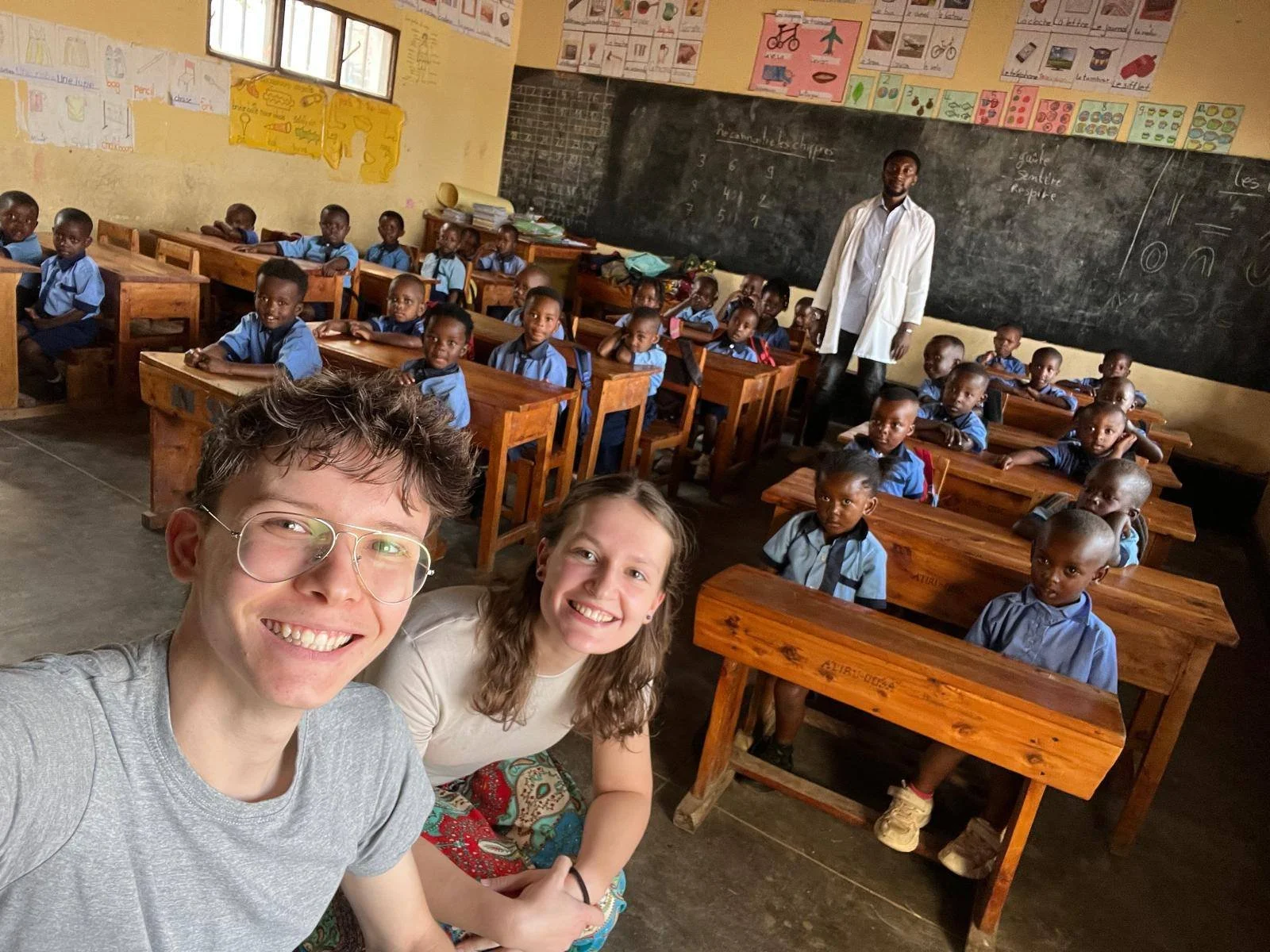 Deux jeunes adultes prennent une selfie dans une classe remplie d'enfants en uniforme scolaire assis à leurs bureaux. Un enseignant se tient devant un tableau noir dans le fond.