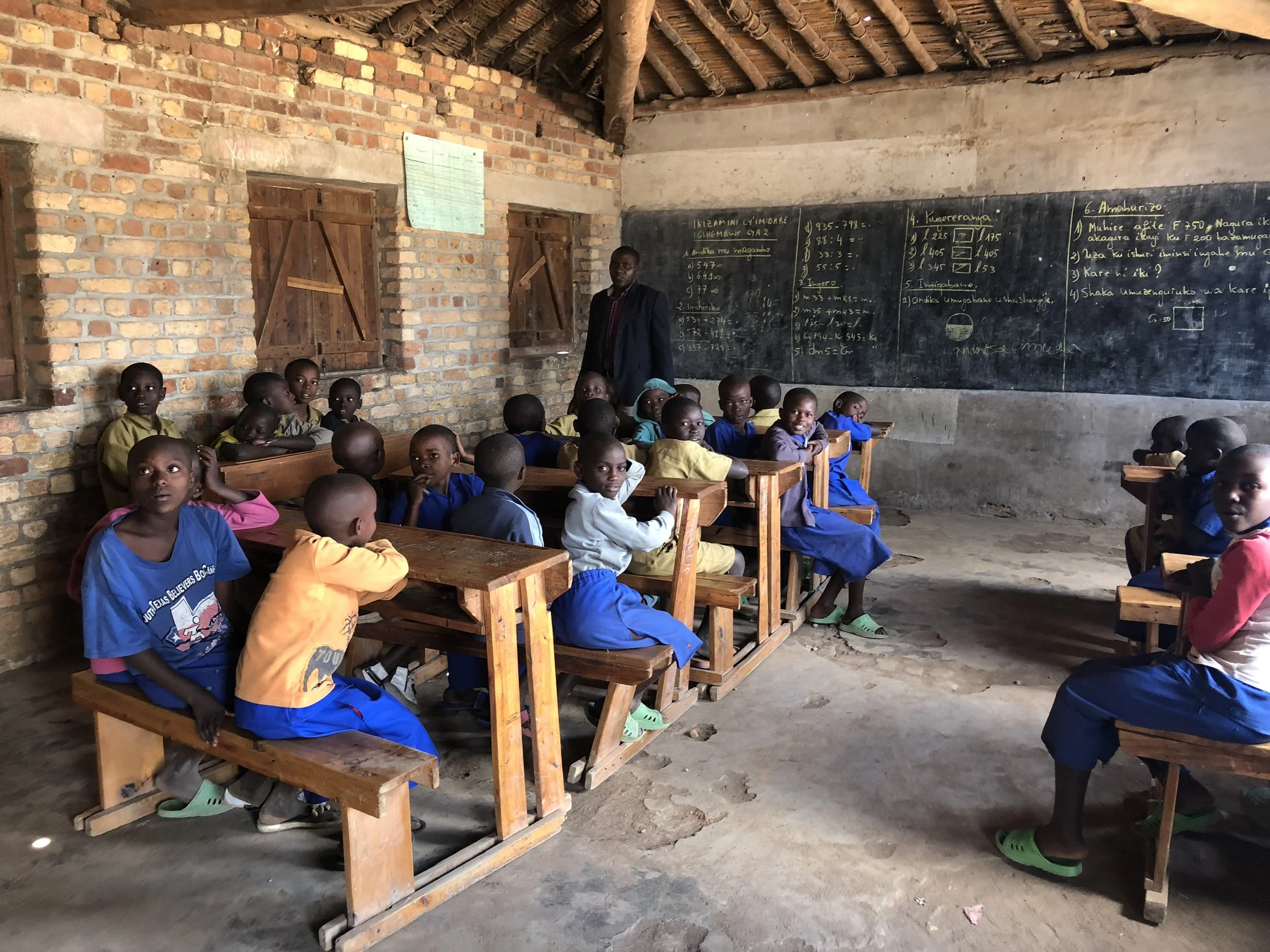 Une classe d'école rurale avec des enfants assis à des bancs en bois, un enseignant à l'arrière, des murs en briques et un tableau noir avec des écritures en langue locale.