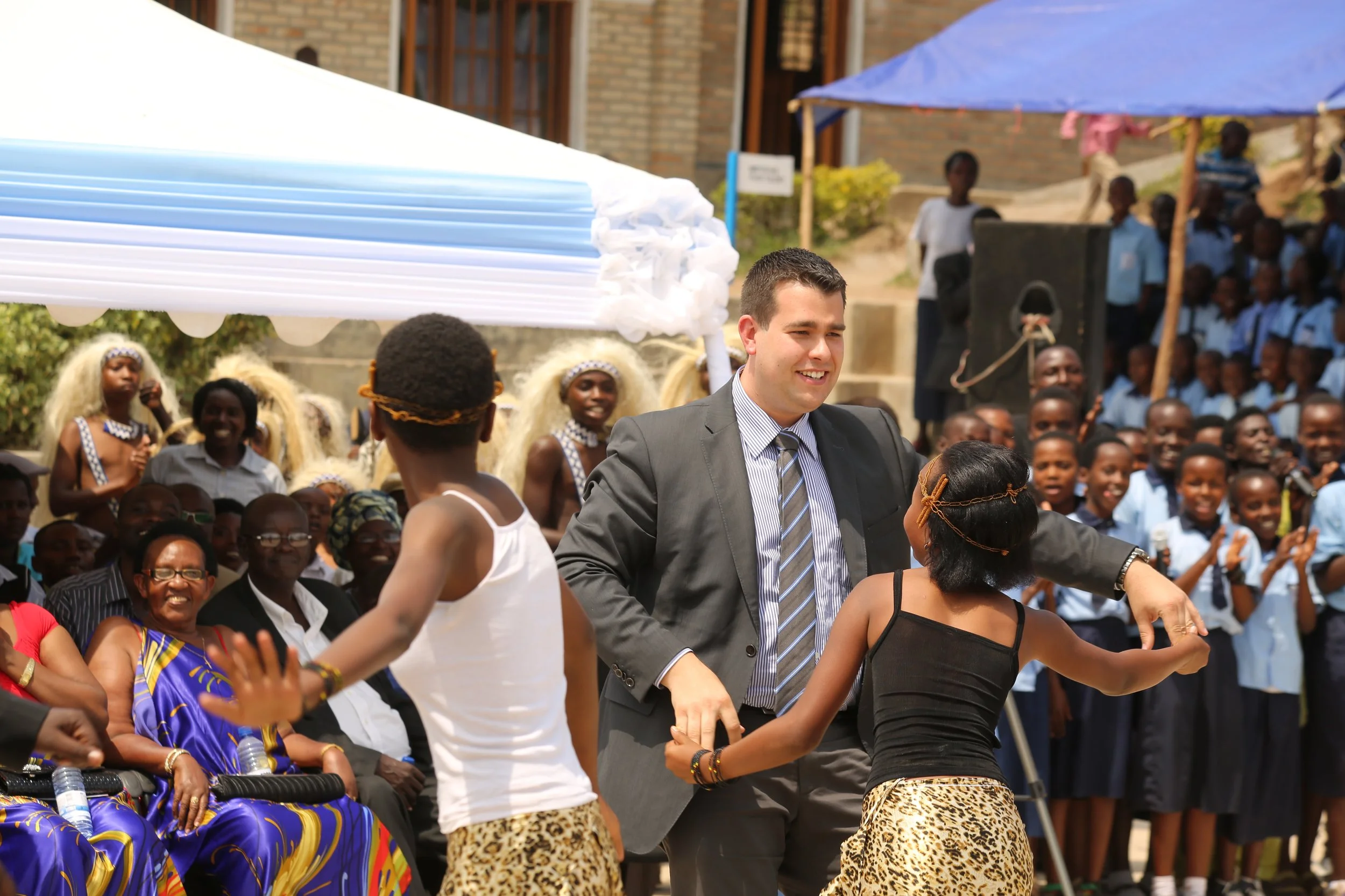 Un homme en costume danse avec deux jeunes filles lors d'un événement en plein air, entouré de spectateurs, principalement des enfants en uniforme scolaire, sous un chapiteau bleu avec d'autres personnes habillées en costumes traditionnels en arrière-plan.