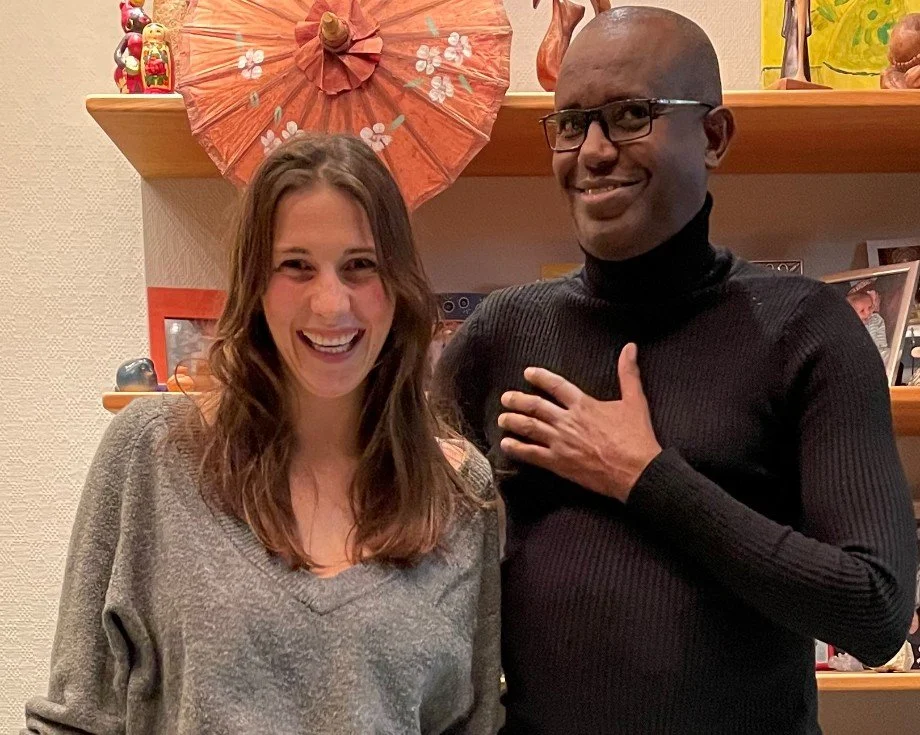 Deux personnes souriantes posent en intérieur, derrière une étagère avec une décoration d'ombre à parasol orange et des cadres photo.