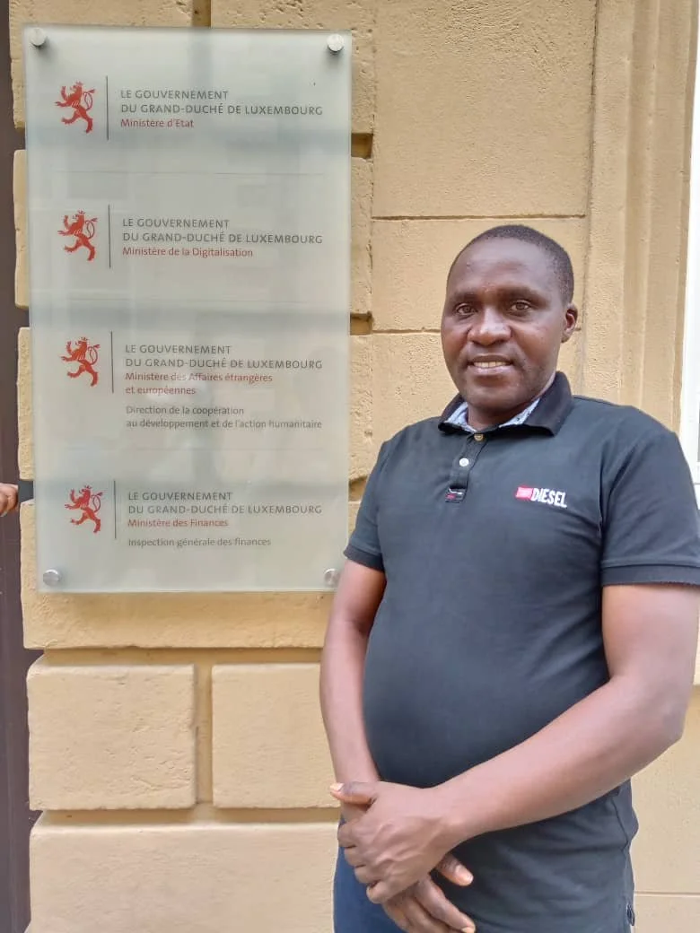 Notre coordinateur de projets posant à côté d'une affiche en pierre avec plusieurs mentions du gouvernement luxembourgeois, y compris le ministère des Affaires étrangères, la Digitalisation, et la Finance, ainsi qu'un logo avec un lion rouge.