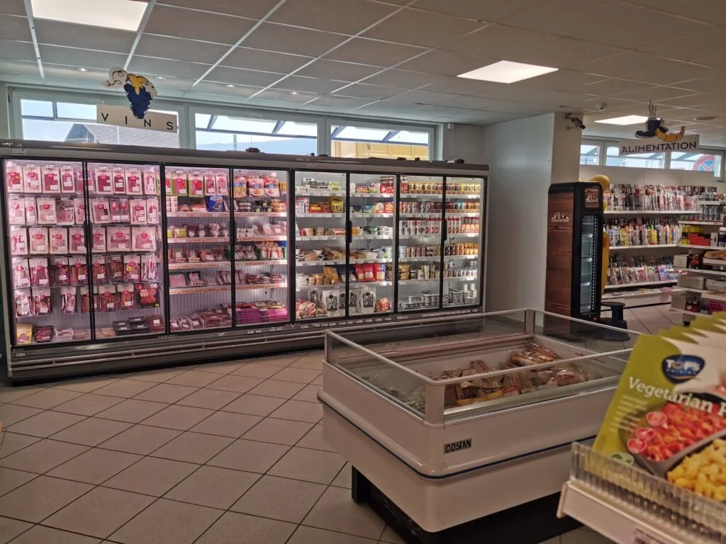 Rayon d'un supermarché avec des produits emballés, notamment des viandes dans un congélateur, et des rayons de livres ou magazines en arrière-plan. Signes indiquant 'VINS' et 'ALIMENTATION'.