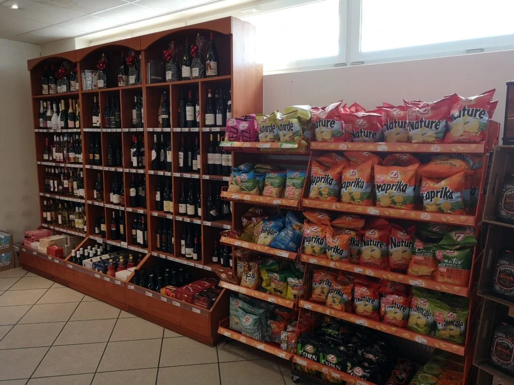Rayon de supermarché avec des bouteilles de vin sur une étagère en bois et des sacs de chips sur une étagère voisine, devant une fenêtre
