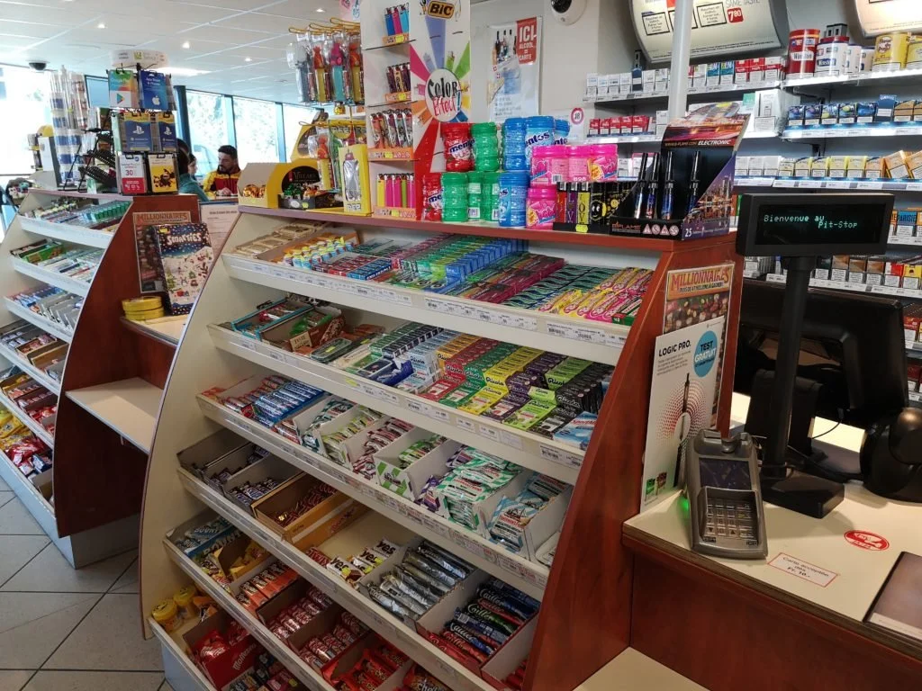 Une étagère dans une pharmacie ou une supérette avec des bonbons, chewing-gums, stylos et fournitures de bureau. Au fond, il y a des rayons avec des médicaments et autres produits.