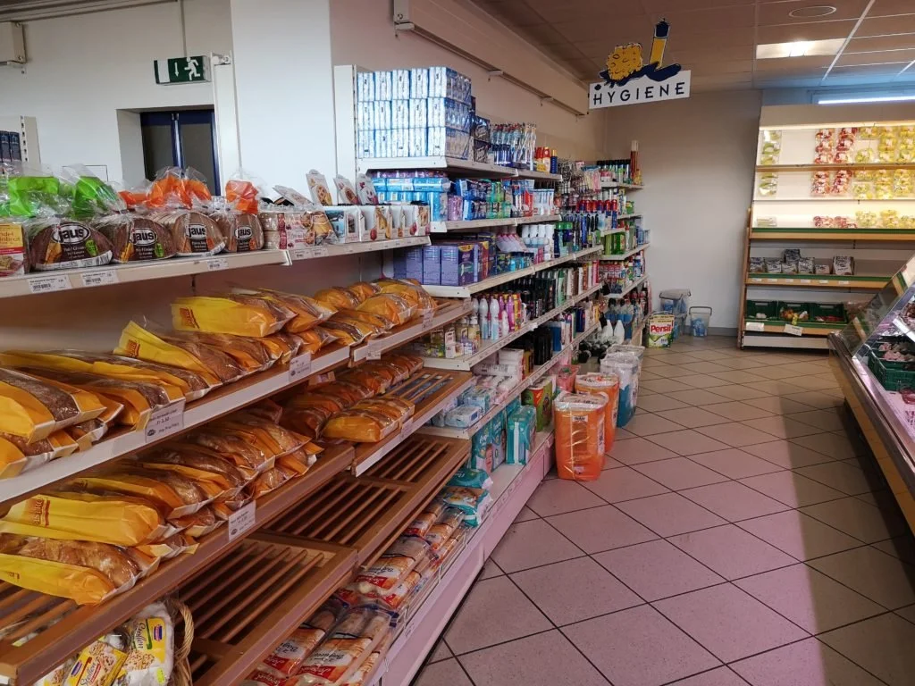 rayon d'une épicerie avec produits alimentaires, notamment du pain en sachet sur la gauche, produits d'hygiène sur la droite, et étagères de produits divers derrière.