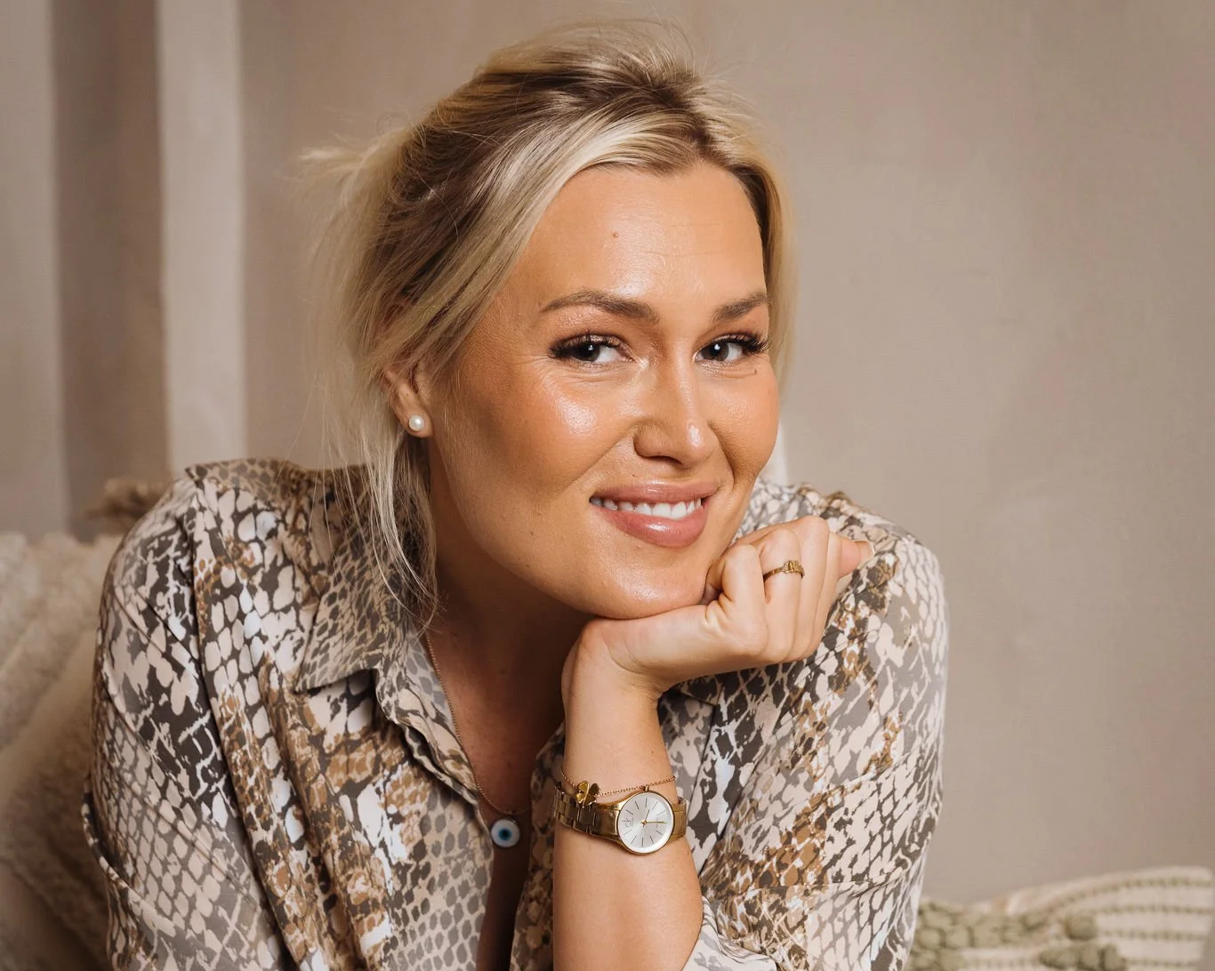 En kvinna med blont hår, leende, bär ett armbandsur och ett par pärlörhängen, poserar med handen under hakan.