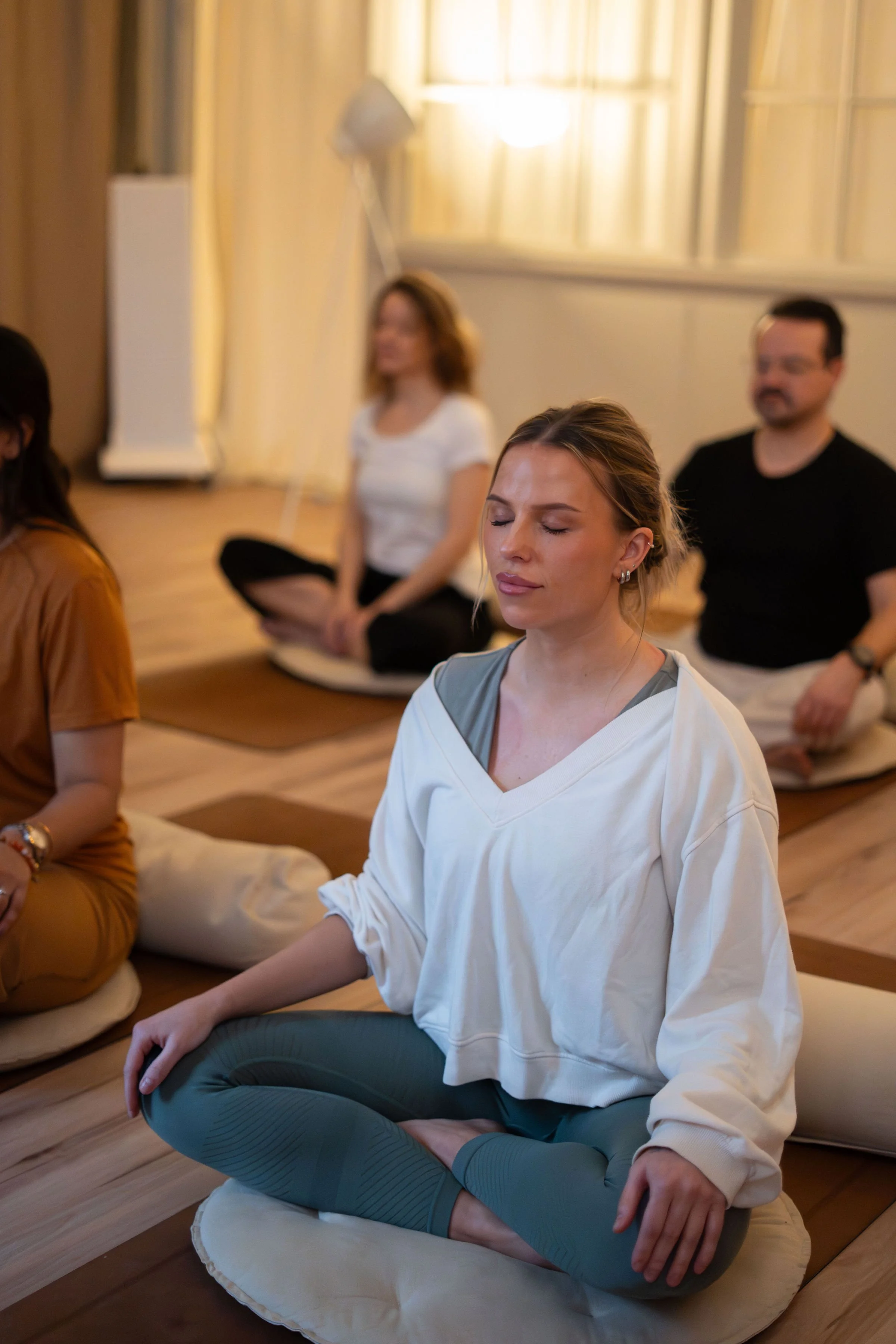 Gruppp med personer som mediterar i ett rum med dämpad belysning, alla i meditationsposition med korslagda ben och slutna ögon.