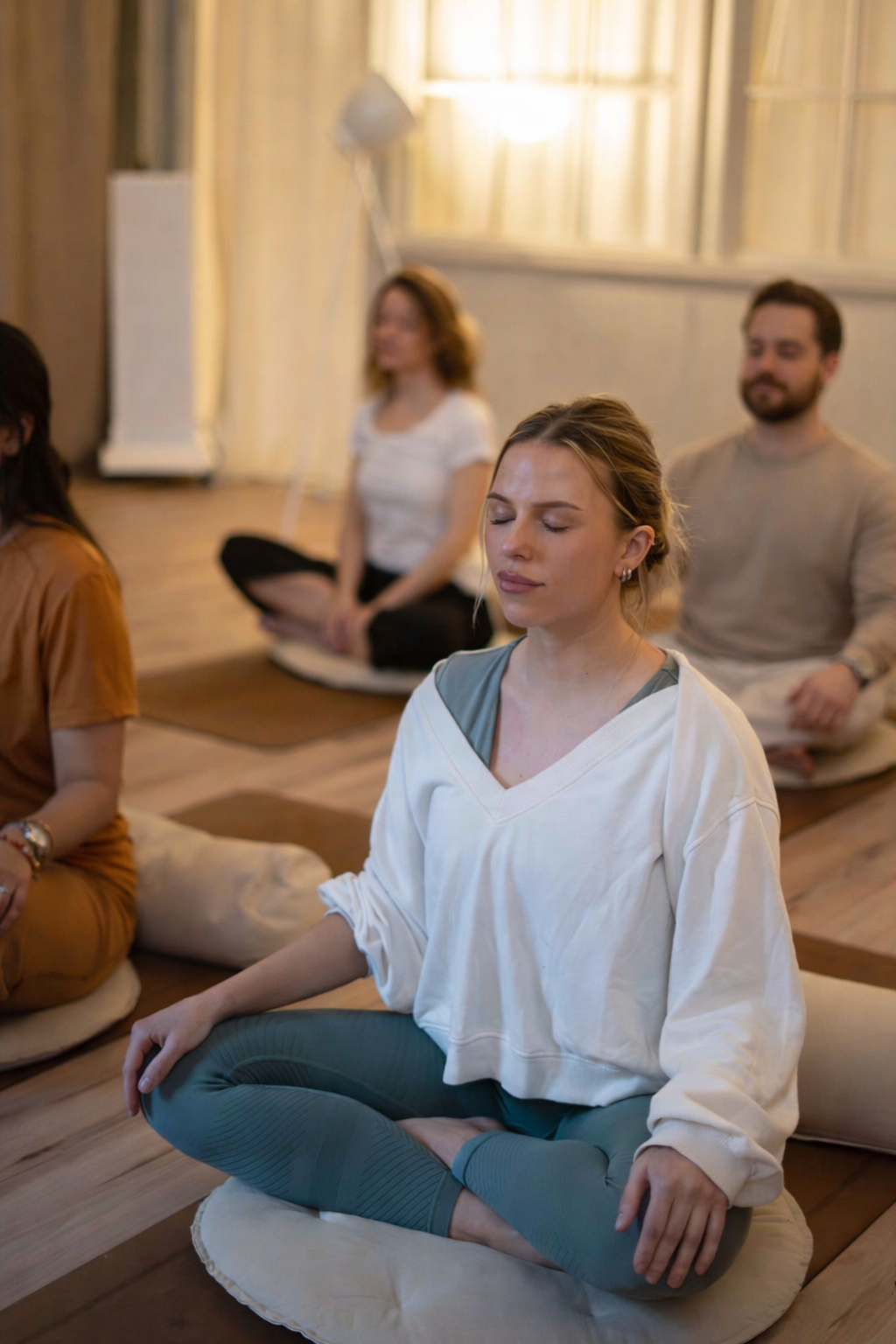 Människor som mediterar i ett rum med mjuk belysning, sittande på golvet med korslagda ben på meditationskuddar.