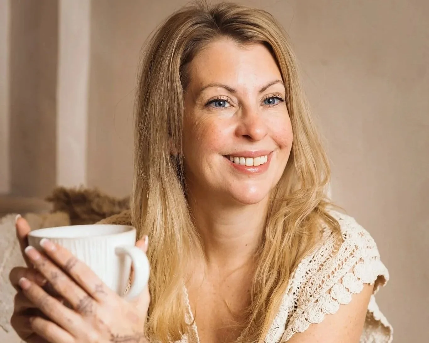 En glad kvinna med långt blont hår, som håller en vit kaffekopp och ler.