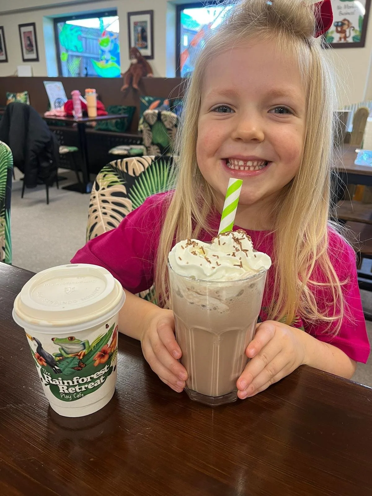 A young girl with blonde hair and a pink shirt is sitting at a restaurant table, smiling, with a milkshake topped with whipped cream and sprinkles, and a striped straw. There is also a cup with a lid on the table. The background shows a colorful, dec